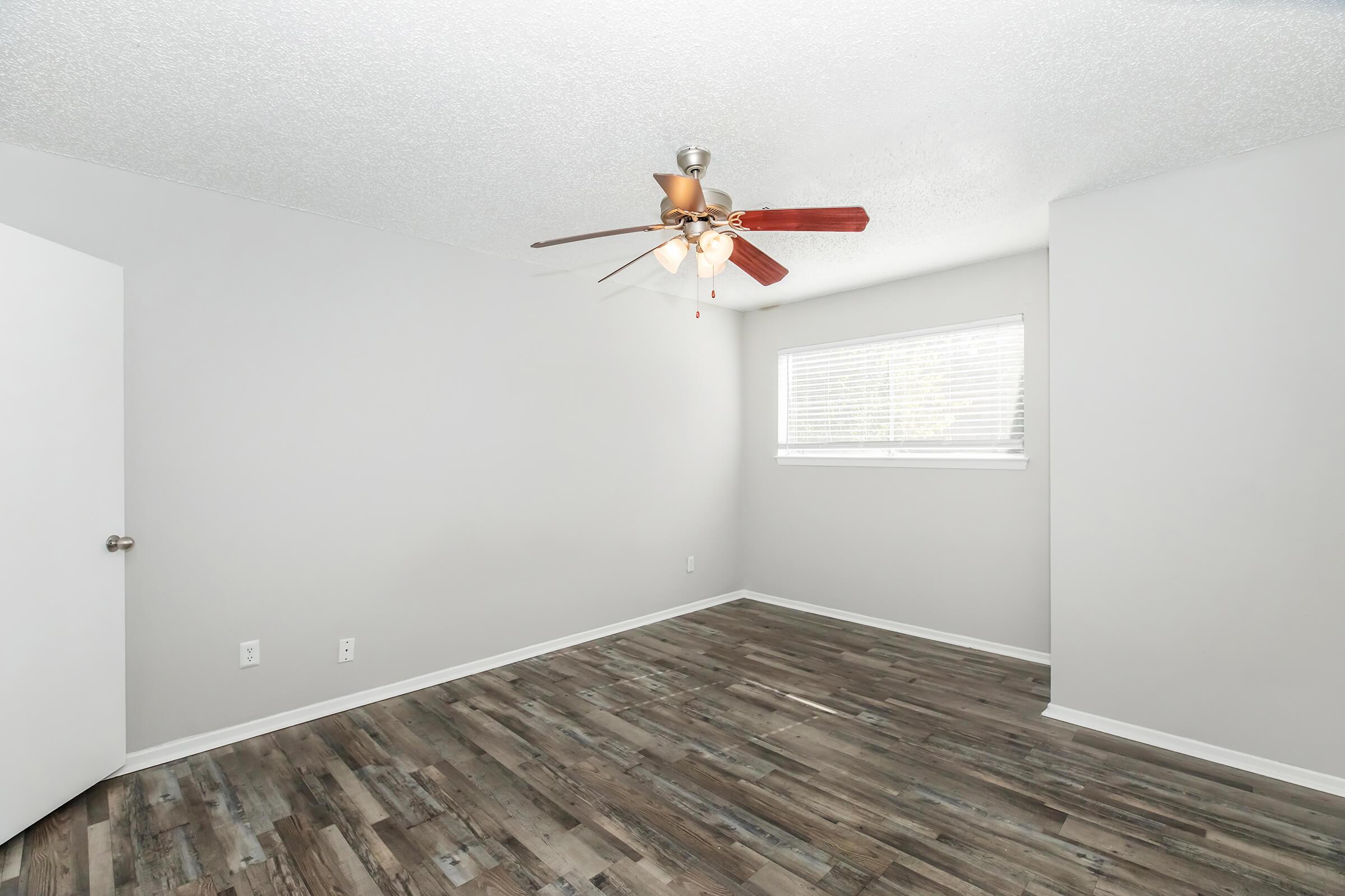 Empty room with light gray walls and a ceiling fan with wooden blades. The floor is covered in wood-like laminate. A window with horizontal blinds allows natural light to enter, and a white door is visible on the left side of the room.