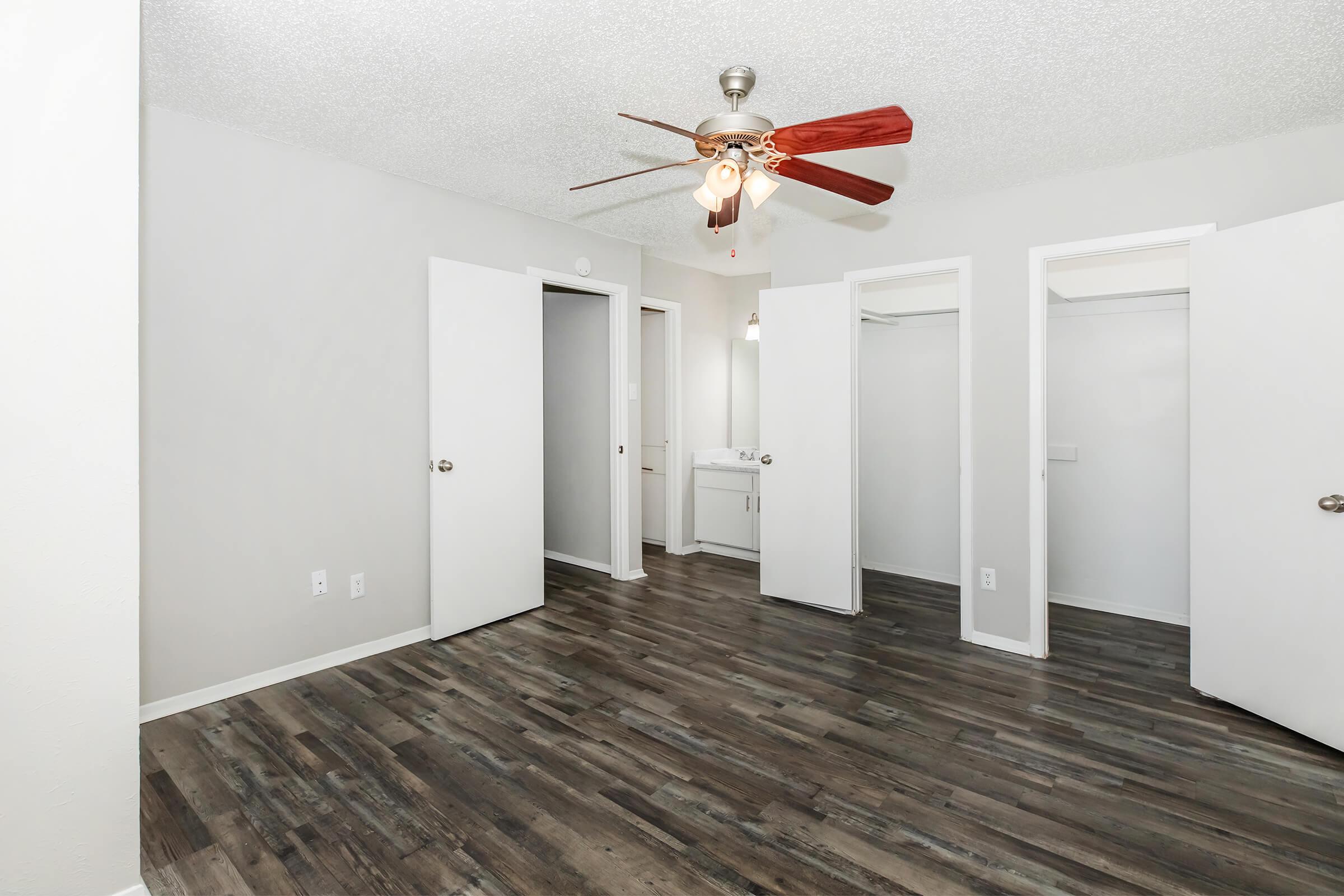Spacious room with a ceiling fan, featuring light-colored walls and wooden flooring. The room includes two open closet doors against the walls and a bathroom area visible in the background. Natural light illuminates the space, creating a bright and airy atmosphere.