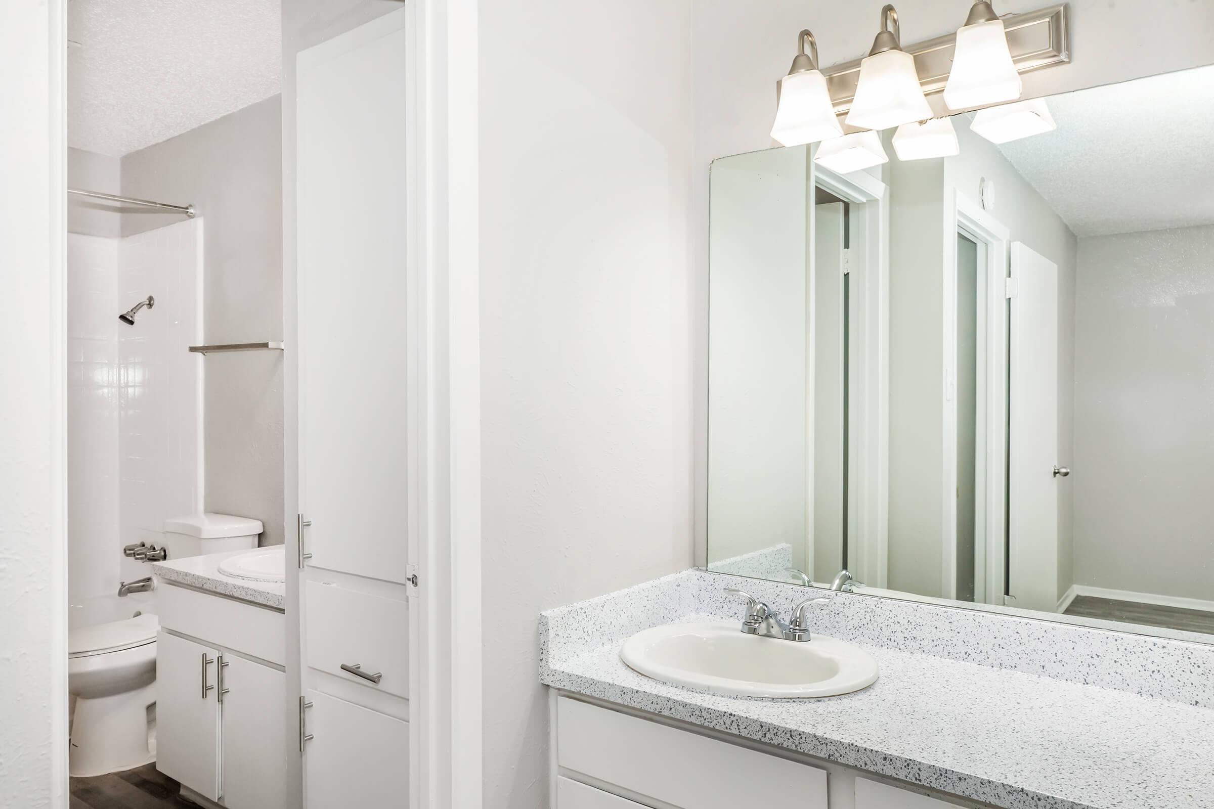 A well-lit bathroom featuring a modern vanity with a white countertop and sink, a large mirror above, and two light fixtures. The background shows a shower area and a toilet behind partially open doors. The walls are painted in a soft gray, enhancing the brightness of the space.