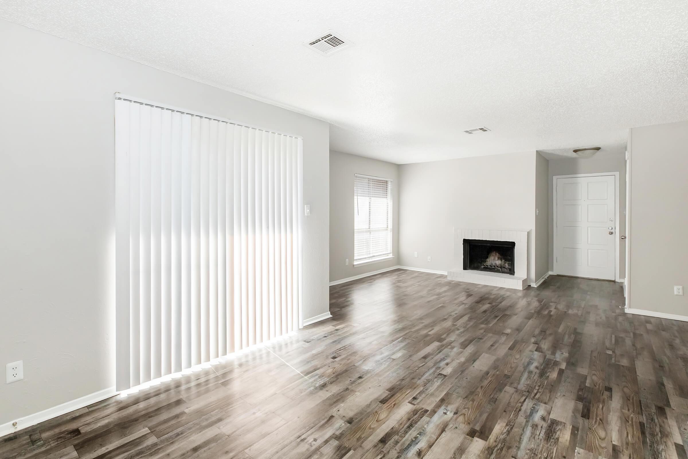 A bright, airy living room featuring hardwood floors, large windows with vertical blinds, a fireplace, and a doorway leading to another room. The walls are painted a light color, contributing to a spacious and inviting atmosphere.