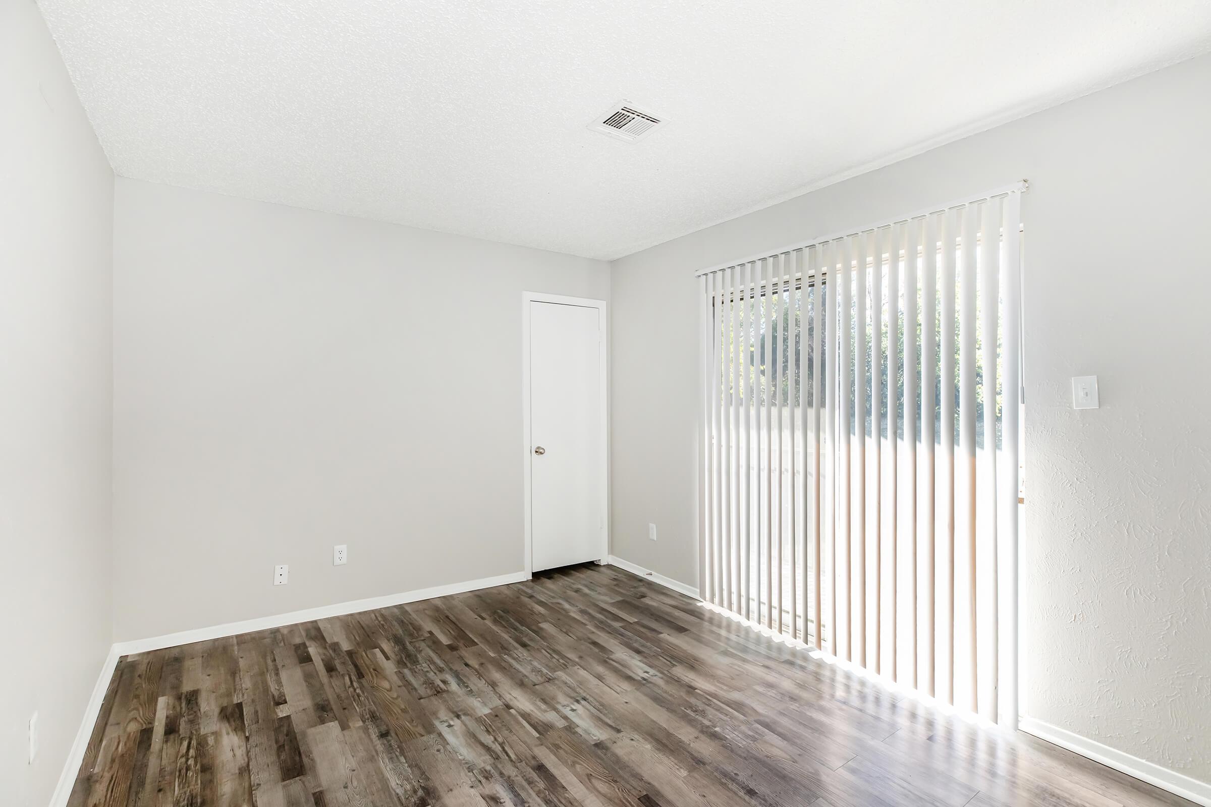 Empty room with light grey walls and wooden laminate flooring. A sliding door with vertical blinds allows natural light to enter. To one side, there is a small closet door. The overall atmosphere is bright and minimalistic.