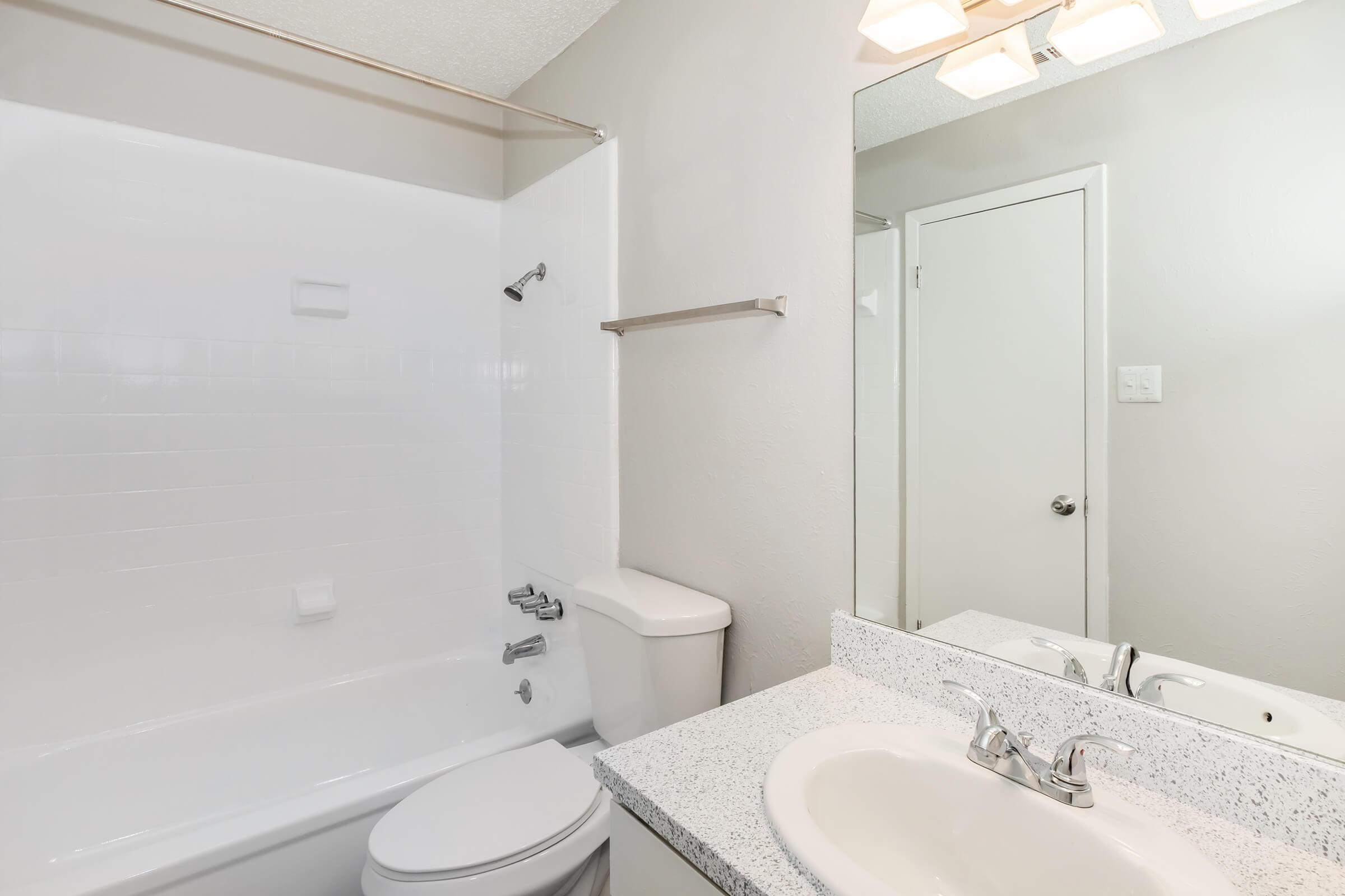 A clean, modern bathroom featuring a white bathtub and shower, a white toilet, and a sink with a granite countertop. The walls are painted light gray, and there is a mirror above the sink. A towel bar is mounted on the wall, and a door leading out of the bathroom is visible.