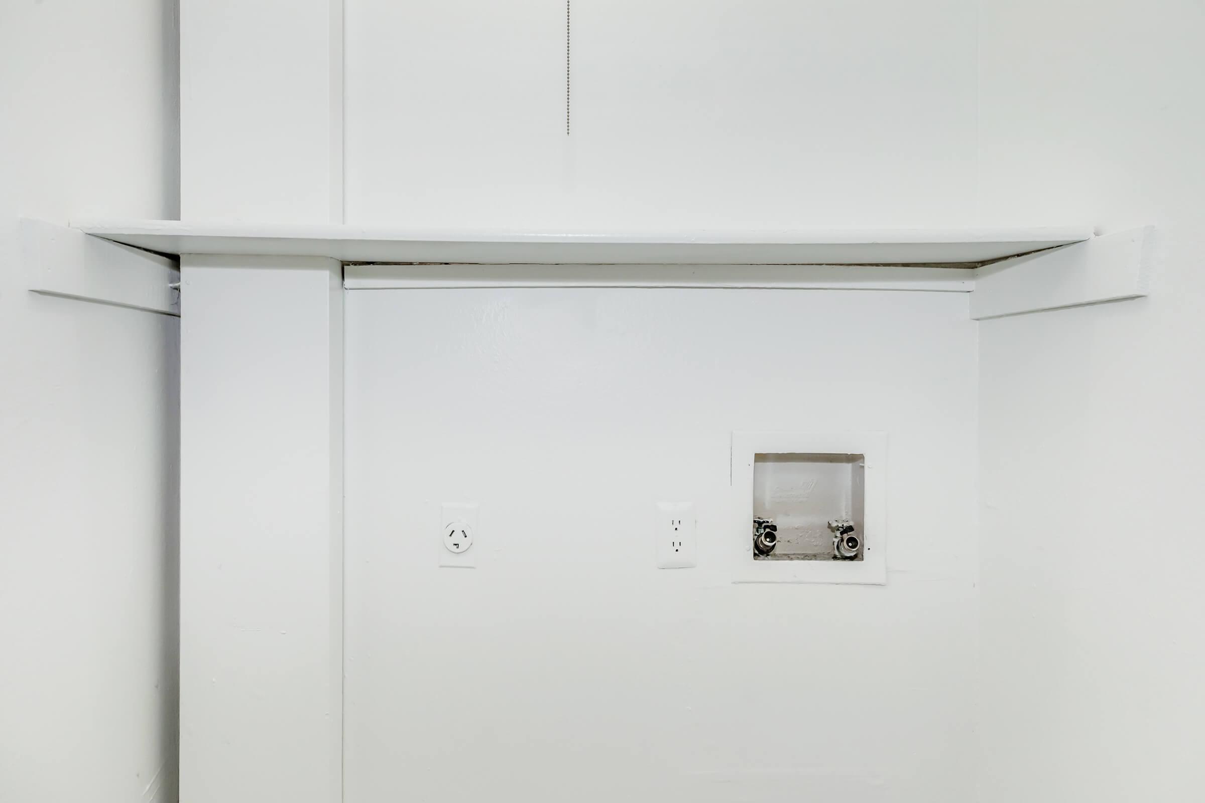 Empty white wall with a horizontal shelf mounted at the top. Below the shelf, there are two electrical outlets on the wall and a small cutout or opening, possibly for a connection or vent. The overall appearance is minimalistic and modern.