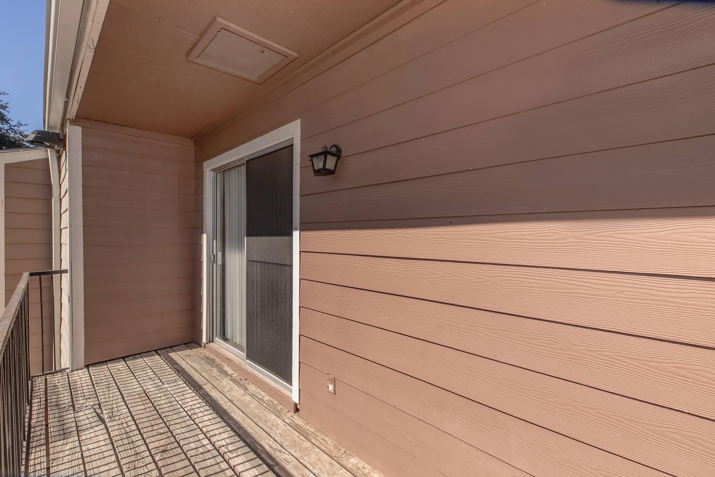A small balcony featuring a beige exterior wall, a sliding glass door with a screen, and a light fixture. The balcony has a metal railing and wooden flooring, creating a simple and understated outdoor space.