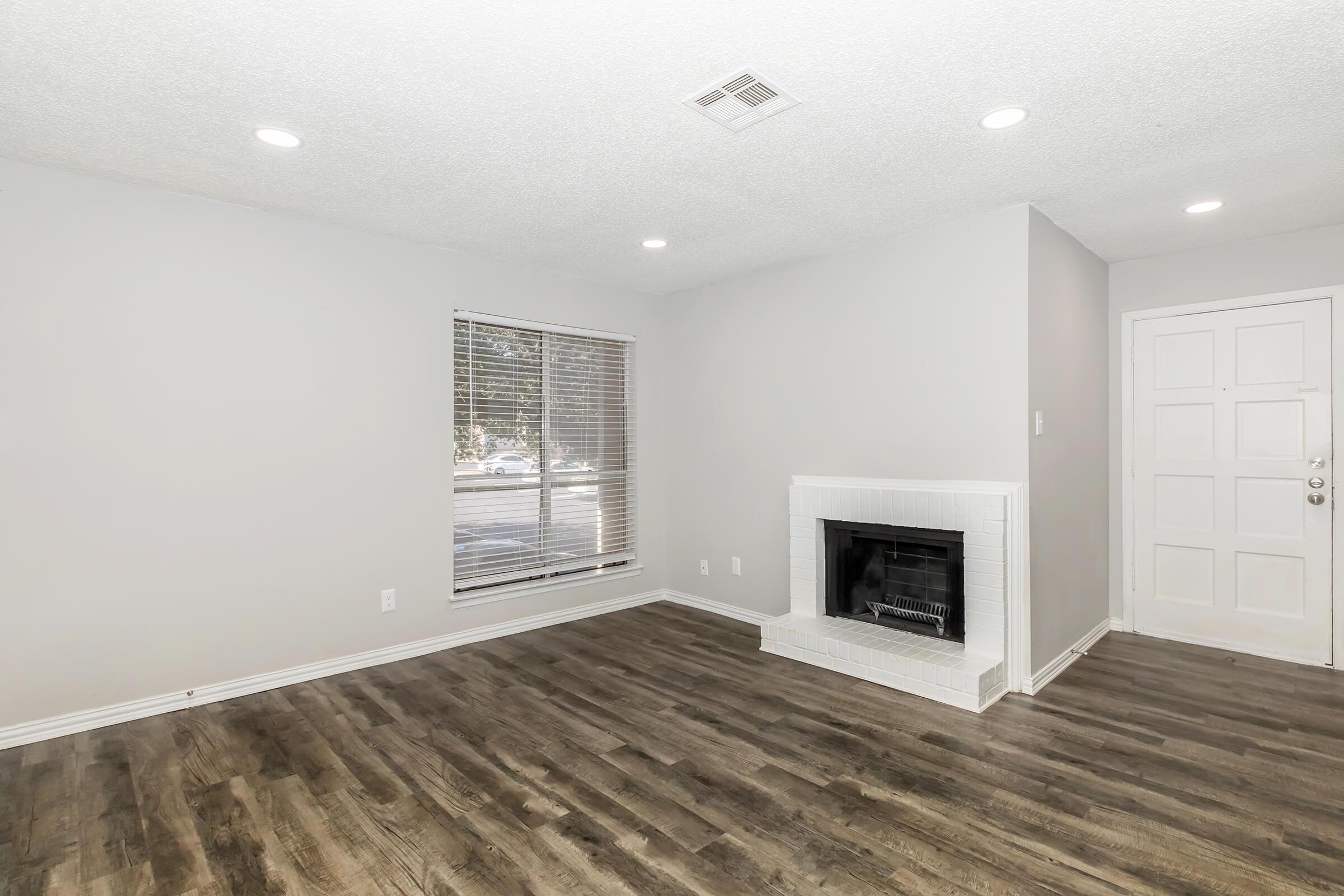 A bright, empty living room featuring a white brick fireplace, large window with blinds, and grey walls. The flooring is wood-like, and there are recessed lights in the ceiling. A door is visible on the right side, leading outside. The space is clean and inviting, ready for furniture and decor.