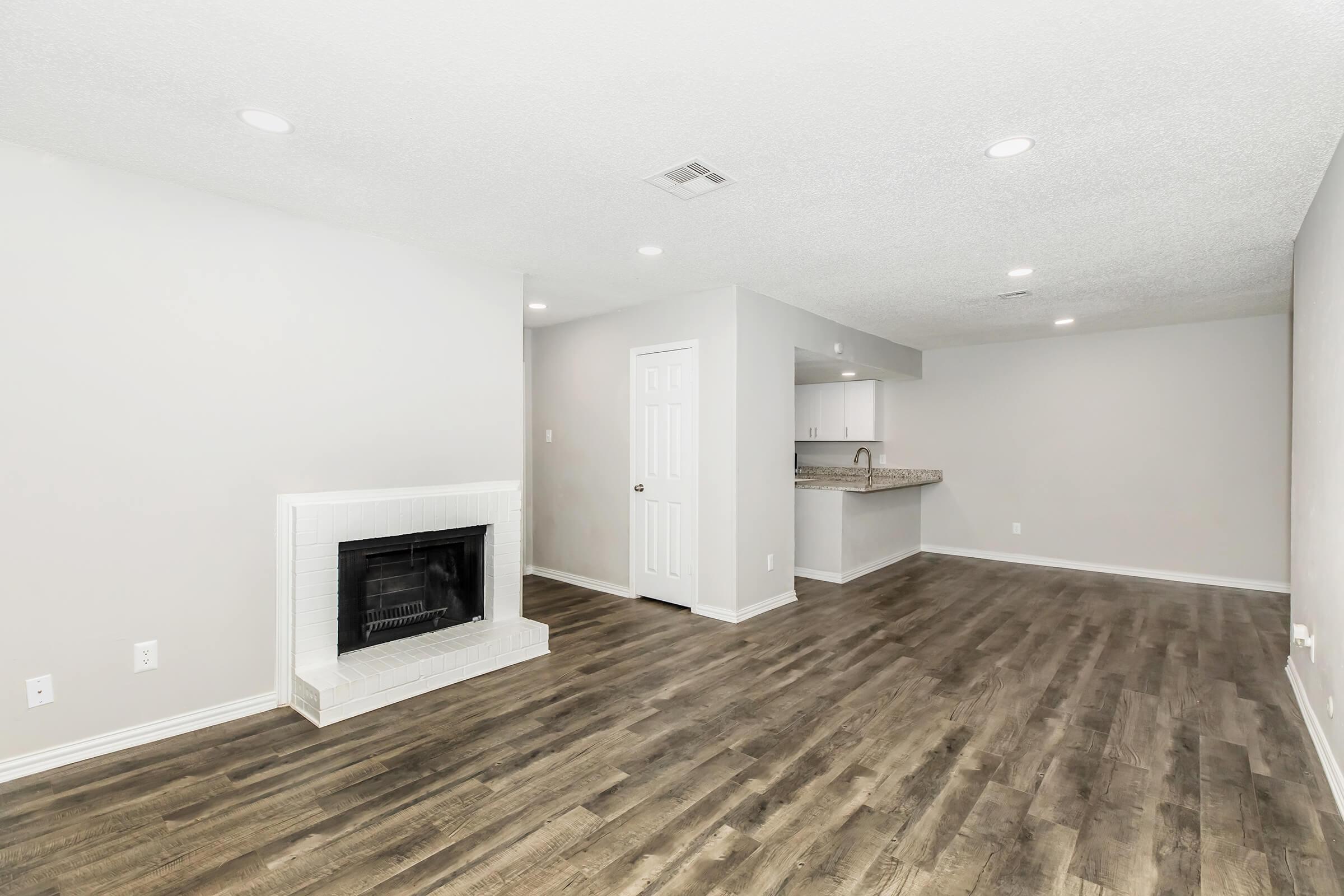 A spacious living area featuring a modern design with light-colored walls, hardwood flooring, and a white fireplace. The room offers natural light and leads to an open kitchen with a countertop. Ceiling lights provide ample illumination, creating a welcoming atmosphere.