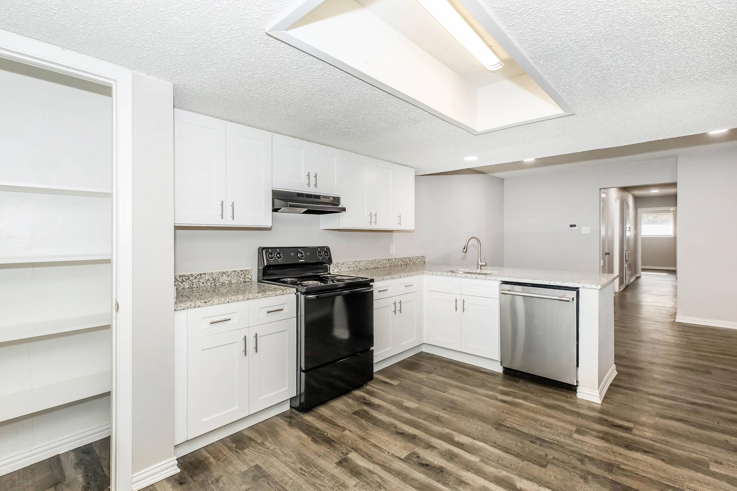 Modern kitchen with white cabinets, granite countertops, and black appliances, including a stovetop and a dishwasher. The space features a skylight for natural light and has wooden flooring. There is an open layout connecting to a living area in the background.