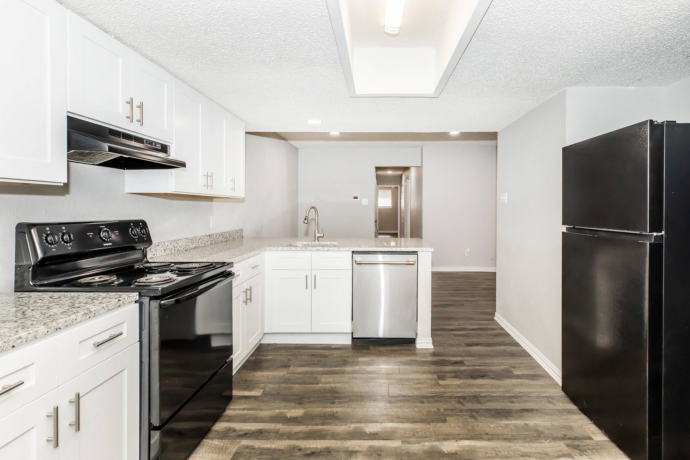 Modern kitchen featuring white cabinetry, granite countertops, a stainless steel sink, and black appliances including an oven, dishwasher, and refrigerator. The space has an open layout with wood-style flooring and well-lit areas, creating a bright and inviting atmosphere.