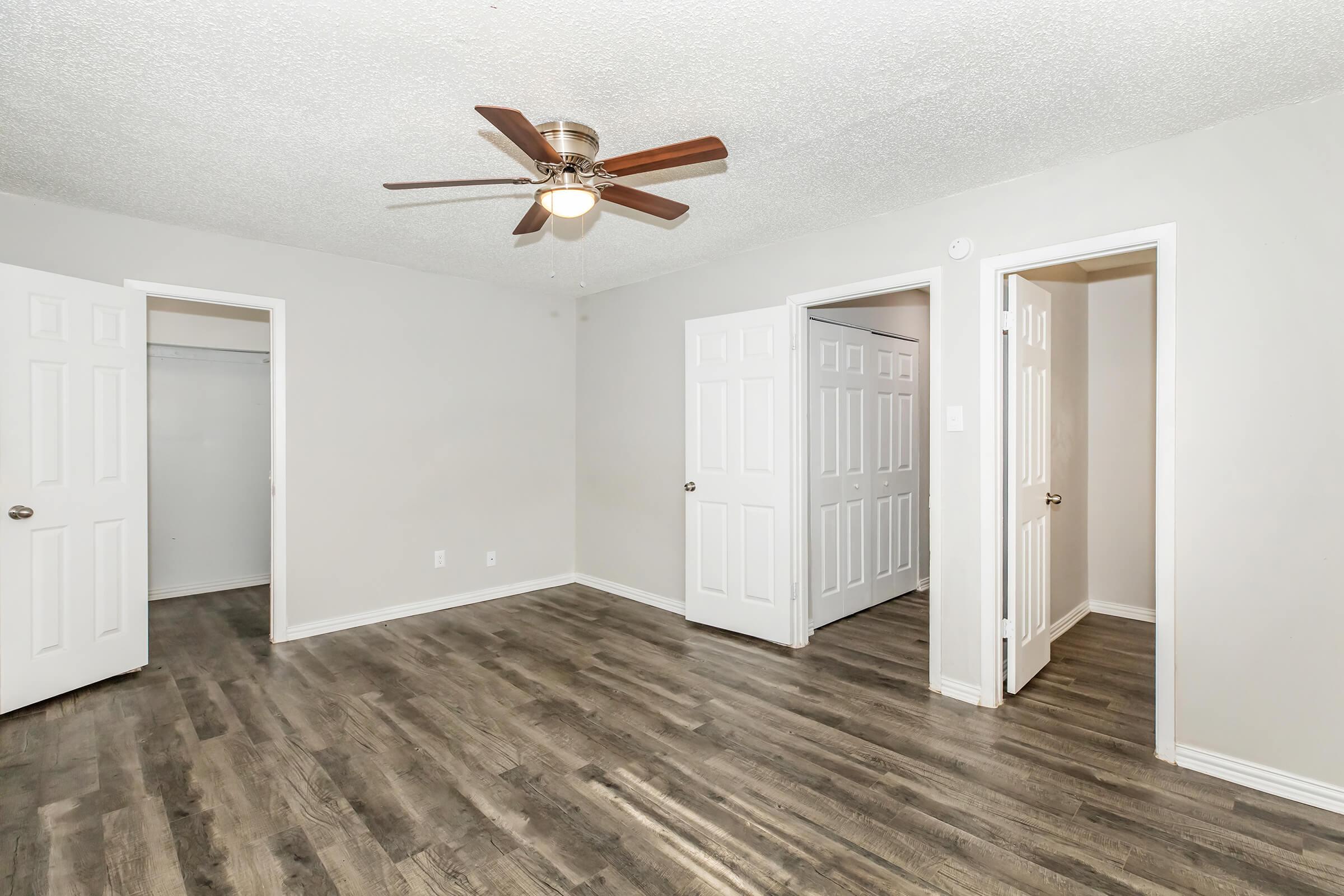 A spacious, well-lit room featuring a ceiling fan, light gray walls, and new laminate flooring. There are two open doors leading to small closets and another room. The space is unfurnished, offering a blank canvas for personal decoration.
