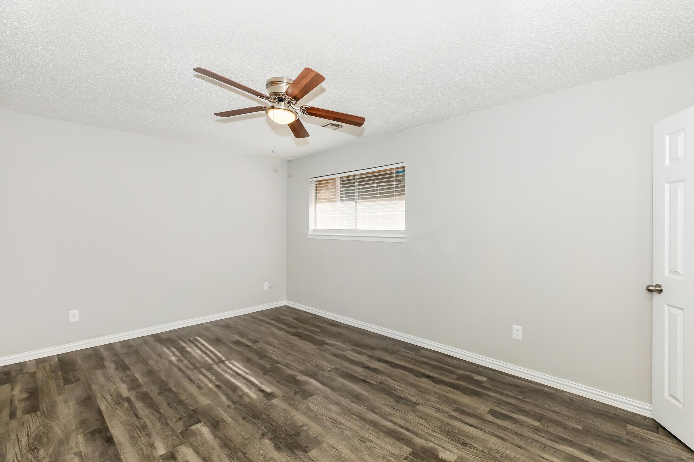 Empty room featuring a ceiling fan, light-colored walls, and hardwood flooring. A window with blinds allows natural light in, and a door is visible on the right side. The space has a modern and minimalist aesthetic, ideal for various uses such as a bedroom or office.