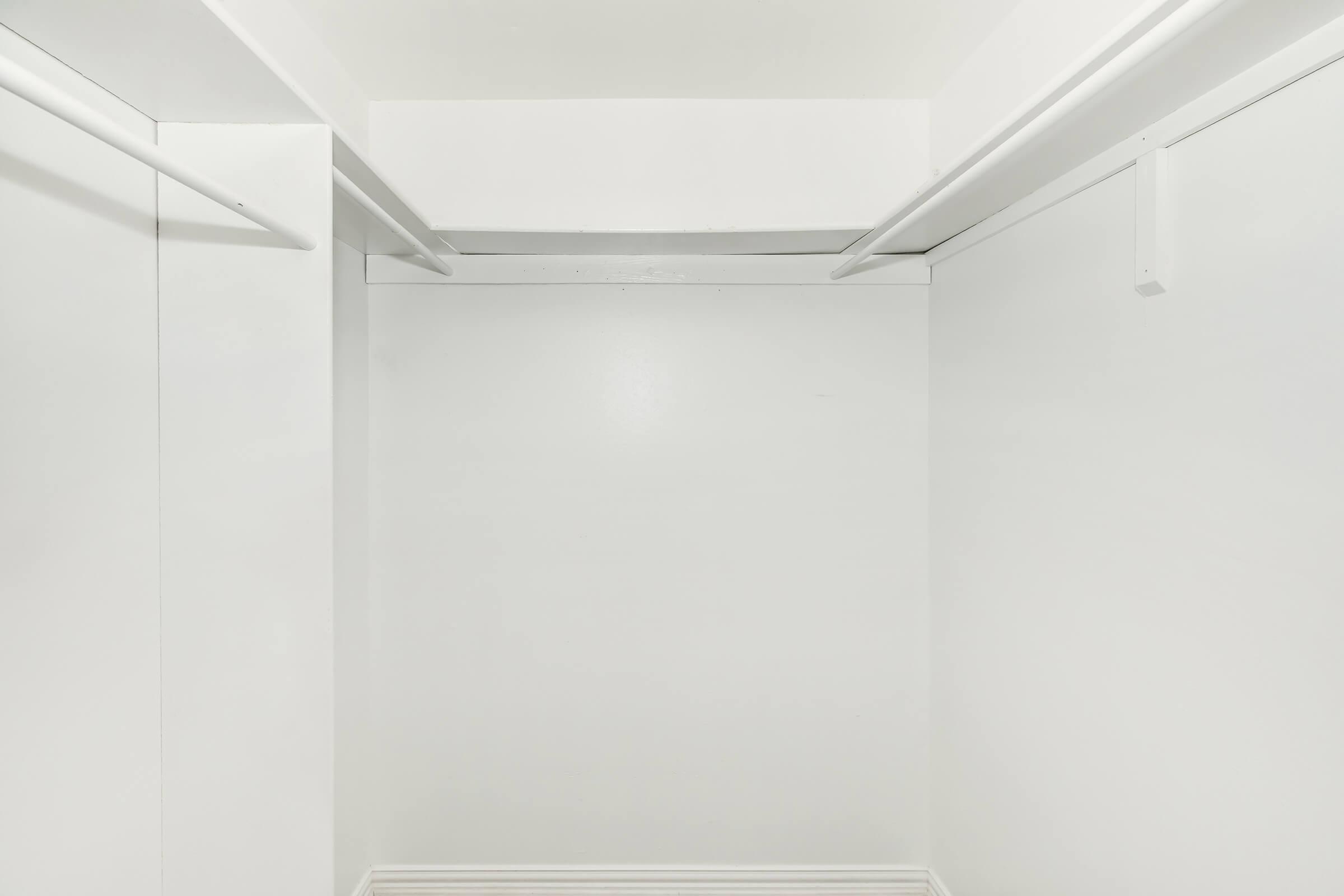 A minimalist, empty closet with white walls and ceiling. Two hanging rods are visible on the upper part of the walls, creating a clean and uncluttered look. The floor is also white, enhancing the overall bright and spacious feel of the interior.