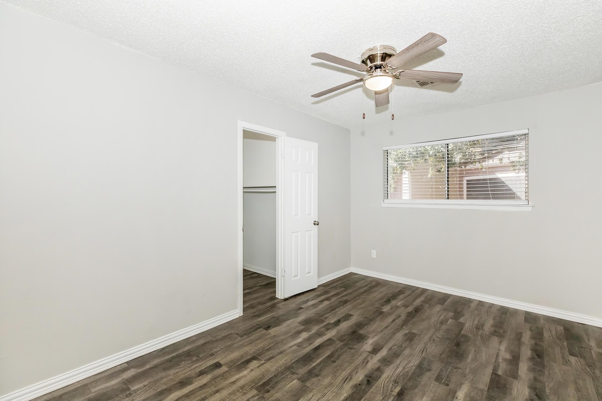 A bright, empty room with a light-colored painted wall and a ceiling fan. It features a window with blinds and a closet with double doors, showcasing wood-like flooring. The space is tidy and ready for furnishing, giving a sense of cleanliness and openness.