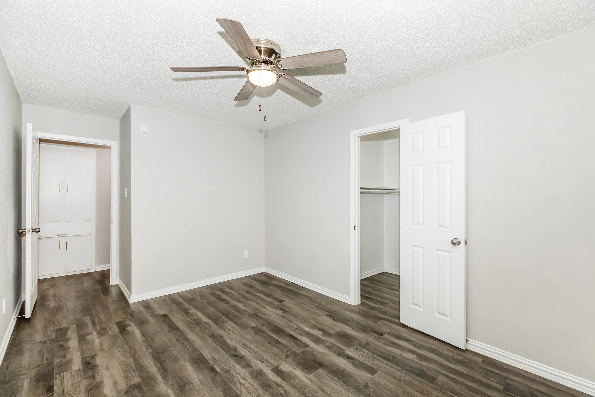 A well-lit room with light gray walls and a ceiling fan. The floor is covered with dark laminate wood. A closet is visible on the left, and a door leads to another area on the right. The overall atmosphere is clean and modern, suitable for various uses.