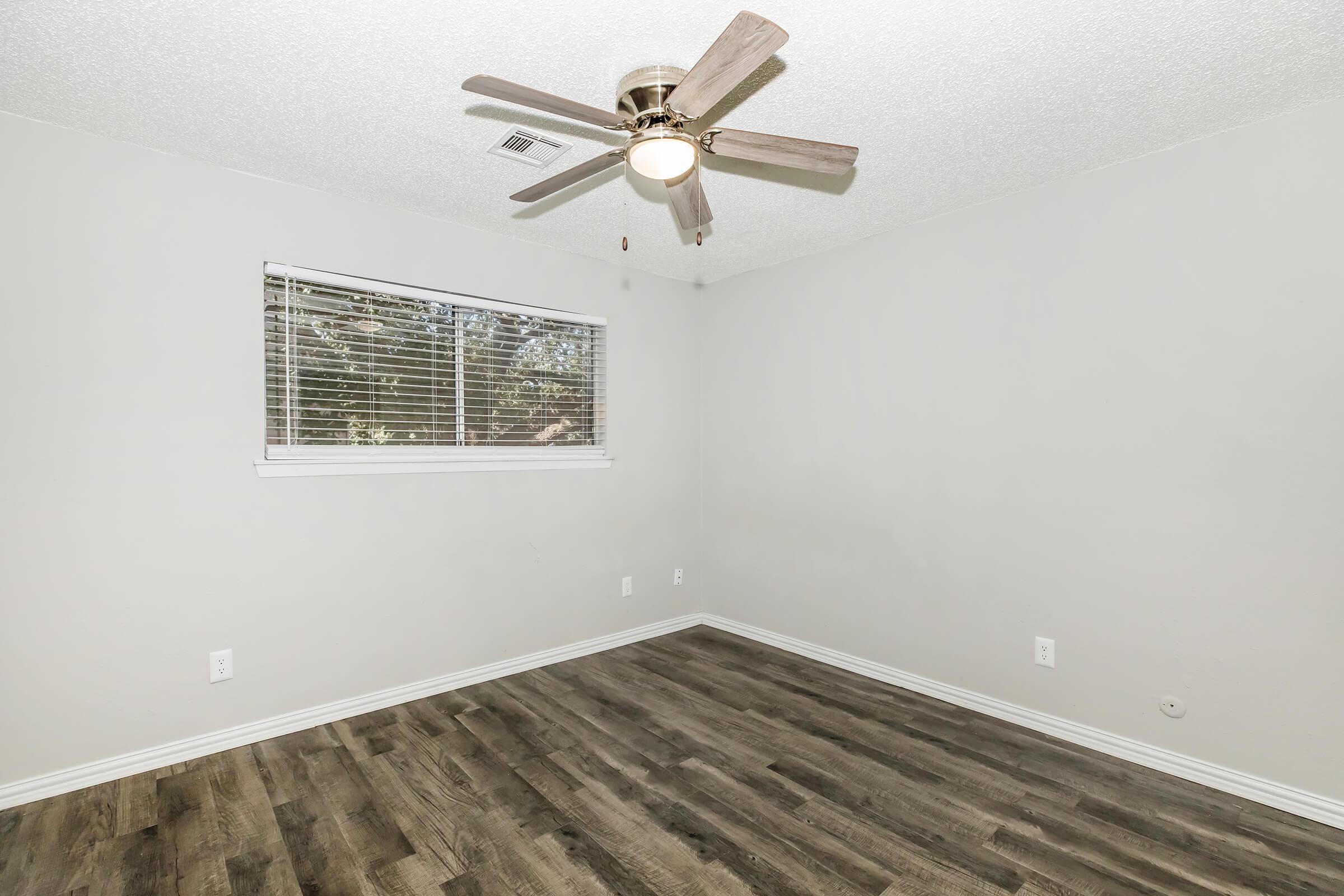 A sparsely furnished, empty room featuring light gray walls, a ceiling fan with wooden blades, and a window with blinds. The flooring is dark wood-like laminate, and there are no decorations or furniture present, making the space appear simple and ready for personal touches.