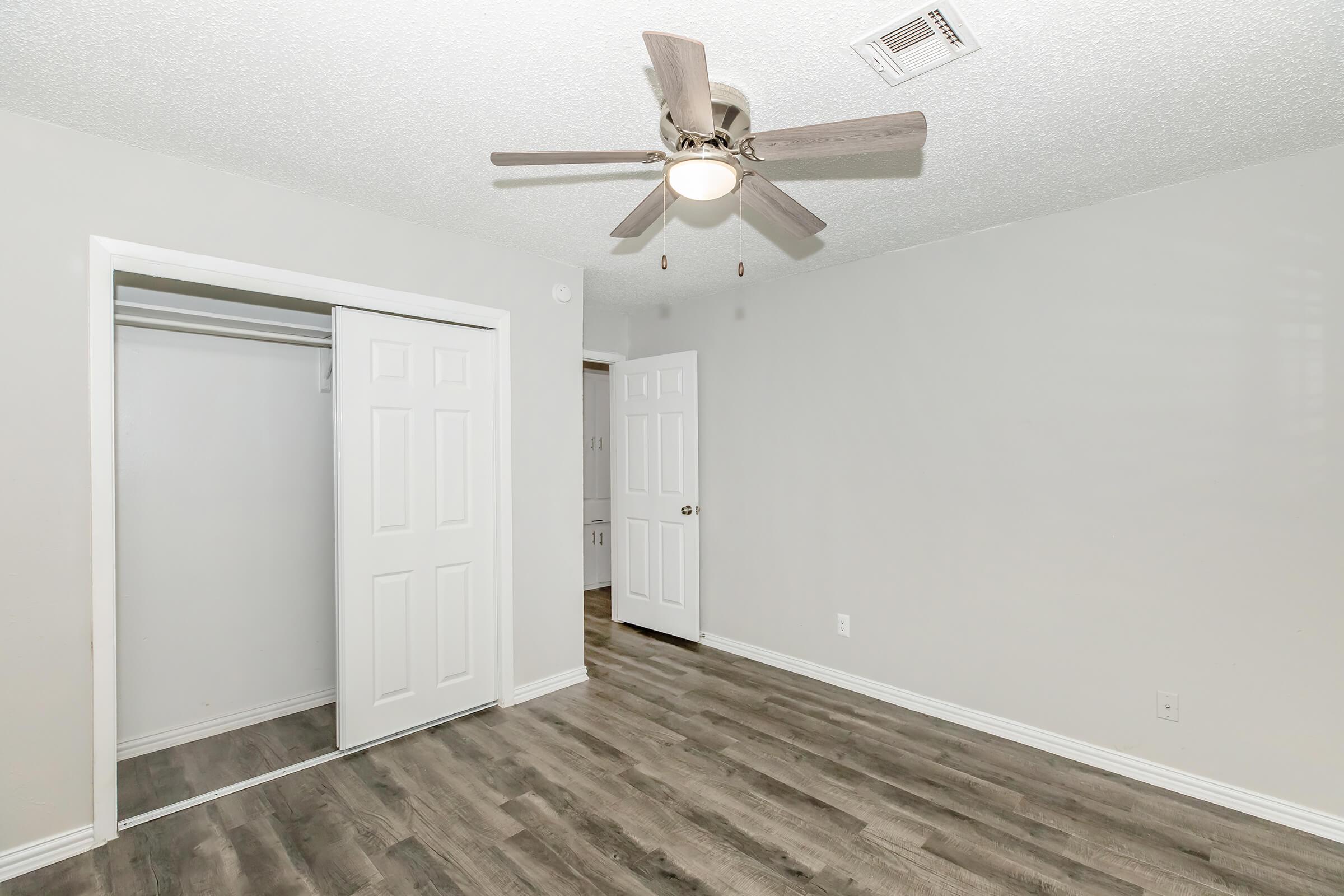A spacious room featuring light gray walls and laminate flooring. The ceiling has a ceiling fan with wooden blades. There is a closet with double doors on one side, and the room opens to another area, suggesting a bright and airy living space.