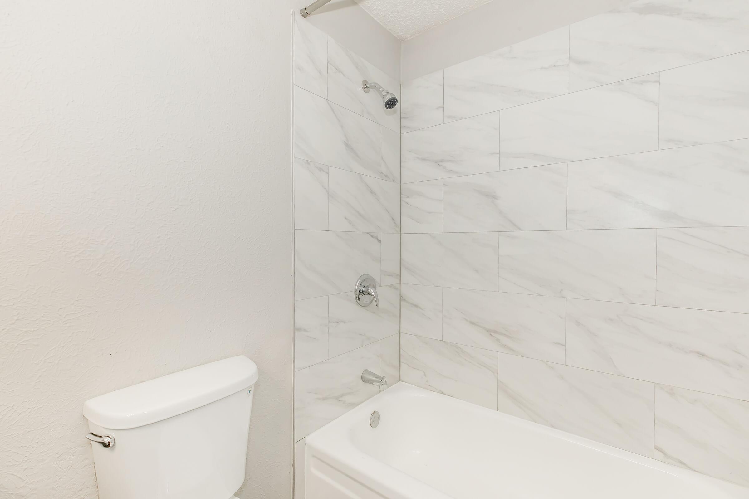 A clean bathroom featuring a white bathtub with a chrome faucet and showerhead, against a backdrop of marble-patterned tiles. To the left, there is a white toilet. The walls are light-colored, contributing to a fresh and modern appearance.
