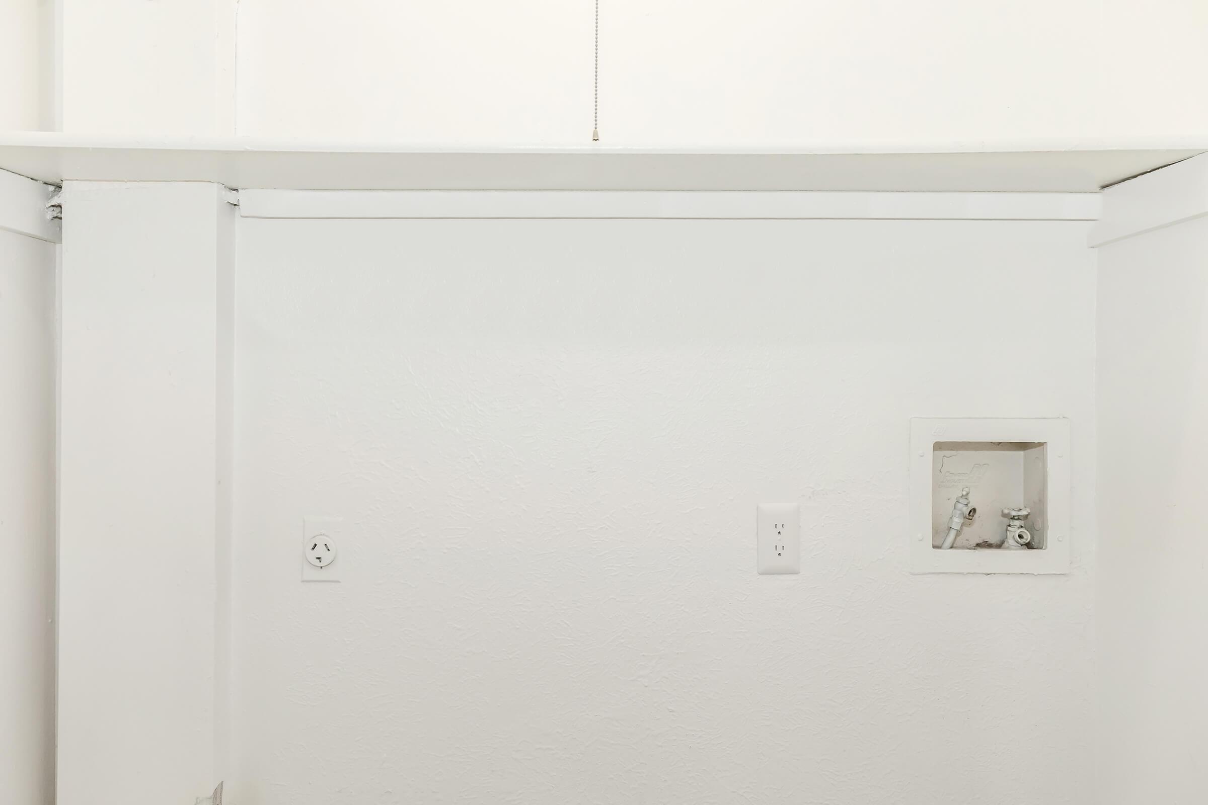 A minimalistic white wall with a small recessed box, likely housing electrical components. There are two electrical outlets visible, one near the floor and the other slightly above it. The overall area appears clean and uncluttered, with natural light illuminating the space.