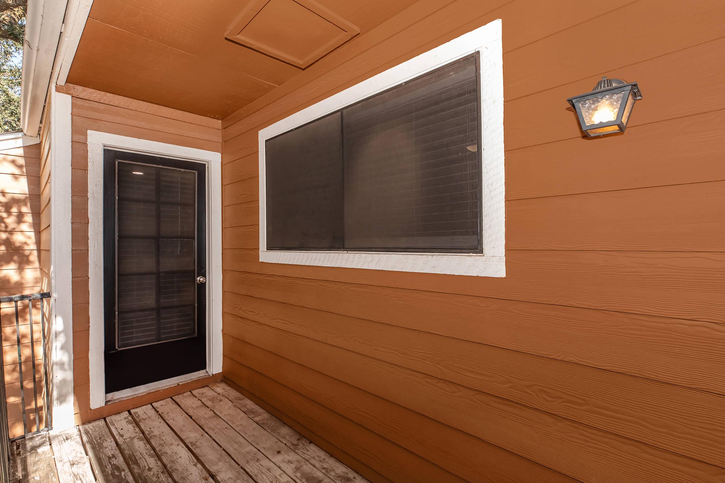A wooden exterior entrance with a dark door and large window. The wall is painted in a warm orange color, and there is a white trim around the window and door. A lantern-style light fixture is mounted near the door, and a wooden deck or floor leads to the entrance, surrounded by natural light.