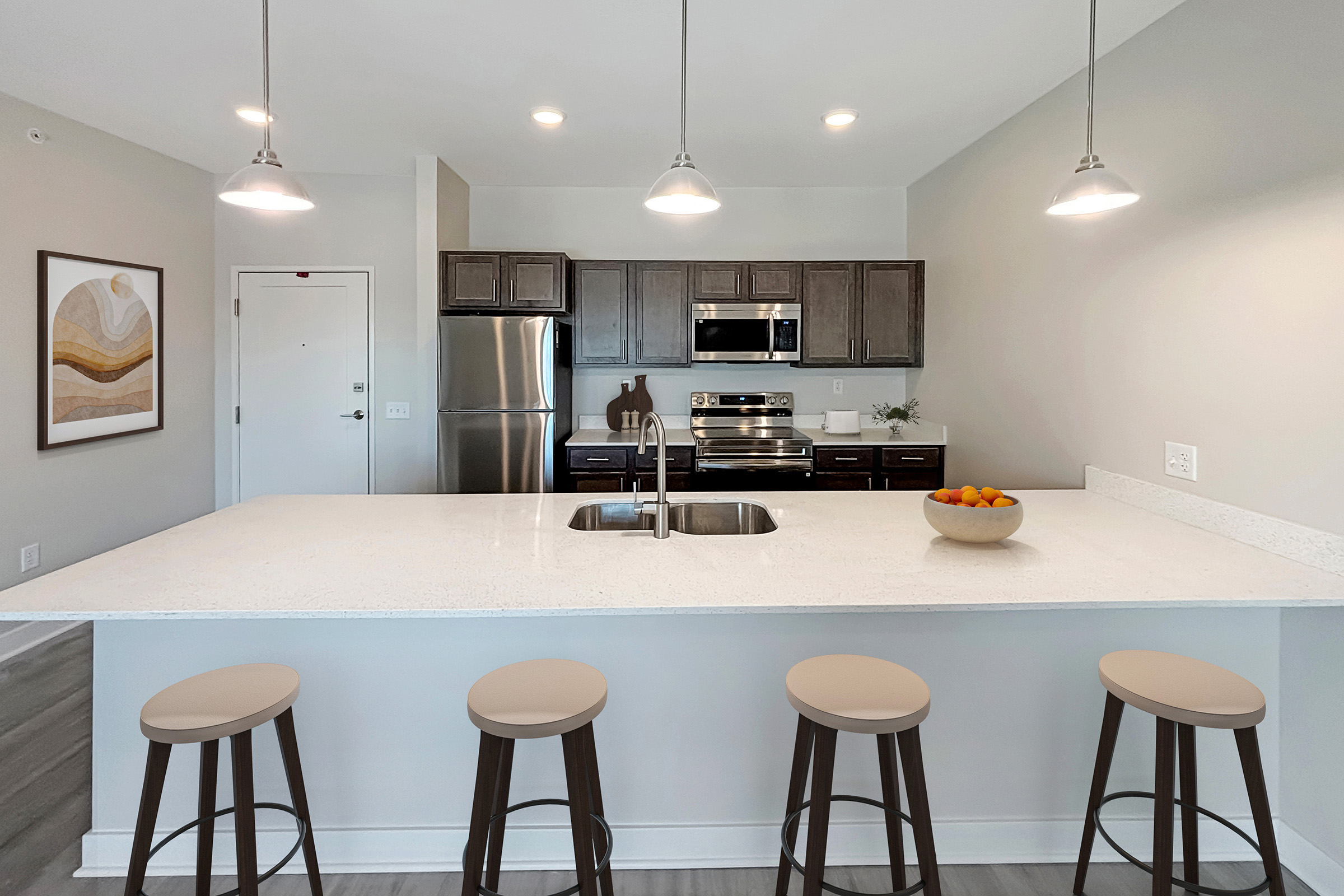 A modern kitchen featuring dark wooden cabinets and stainless steel appliances. The large island has a white countertop with a sink, surrounded by four beige bar stools. Pendant lights hang above the island, and a bowl of oranges sits in the center. The walls are painted light gray, creating a bright and airy atmosphere.