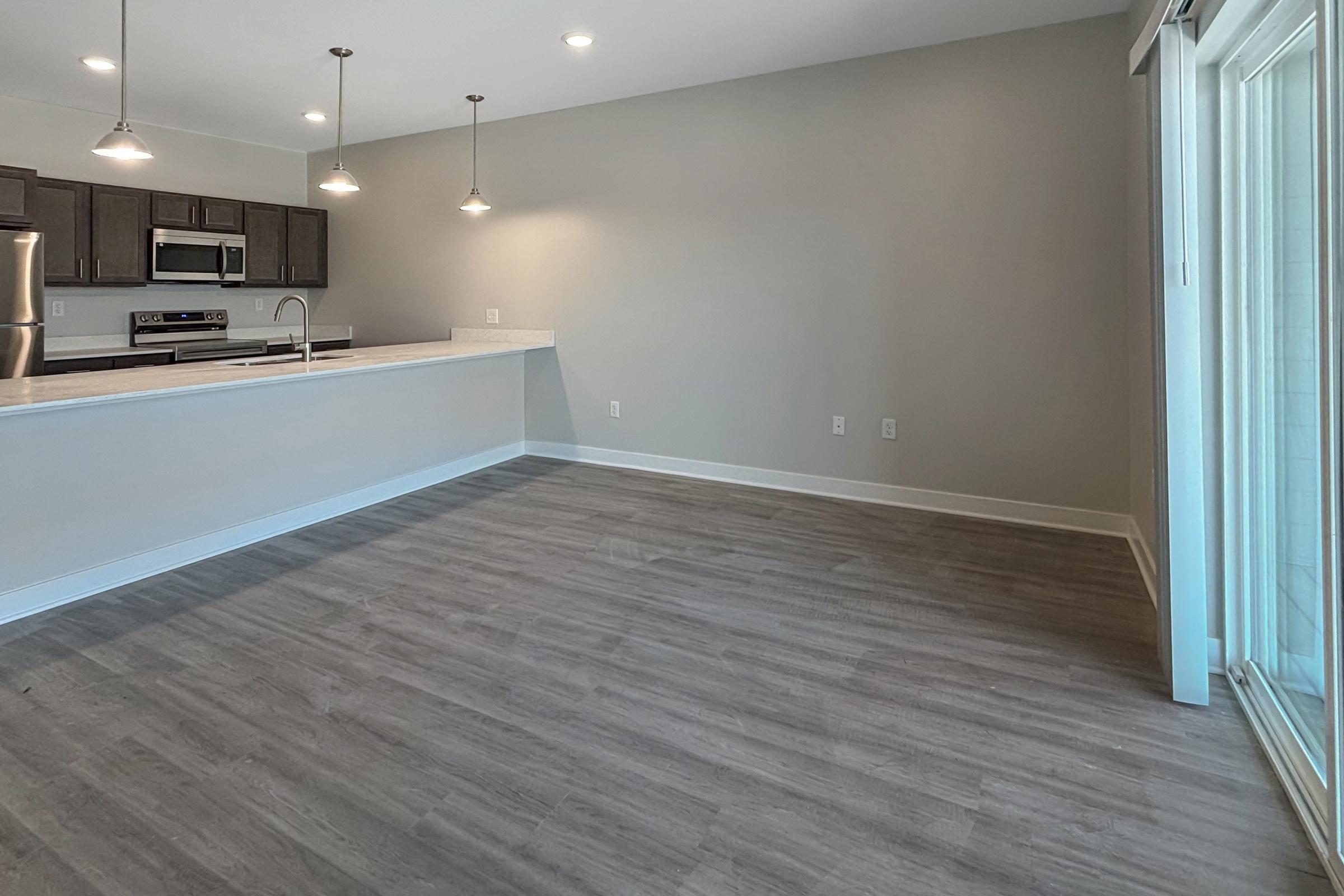 Spacious modern living area with light gray walls and wood-style flooring. The open kitchen features stainless steel appliances, a large countertop, and pendant lighting. There are sliding glass doors leading to a balcony or patio, creating a bright, airy atmosphere.