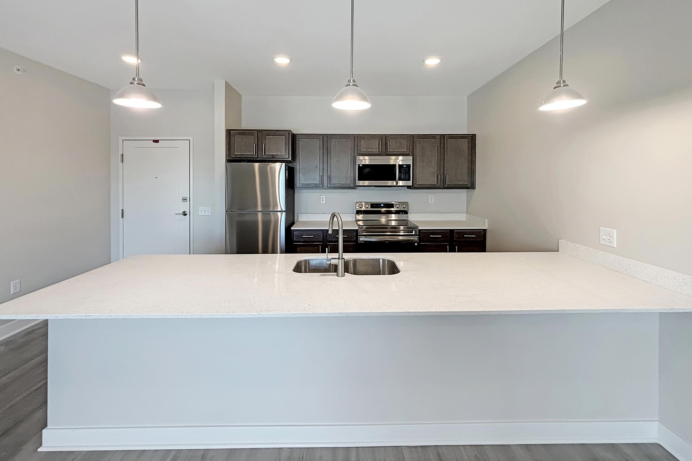 Modern kitchen featuring a large island with a double sink, stainless steel appliances, including a stove and microwave, and dark wood cabinetry. The space is well-lit with pendant lights and has a light, neutral color scheme. A doorway can be seen in the background.