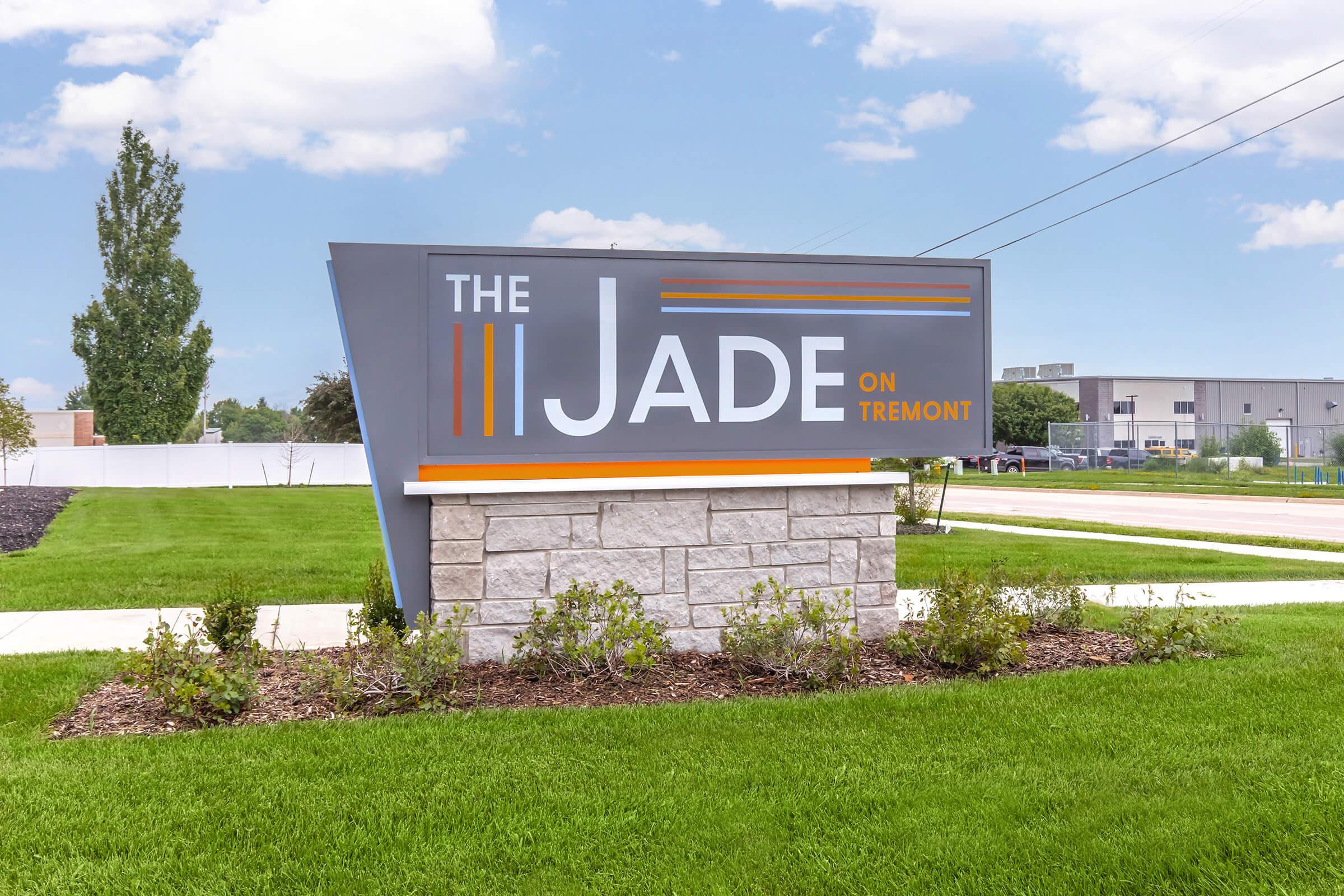 Sign for "The Jade on Tremont," featuring modern design with gray and orange colors. The sign is situated on a green lawn, with shrubs in front and a clear sky in the background. The setting appears tidy and well-maintained.