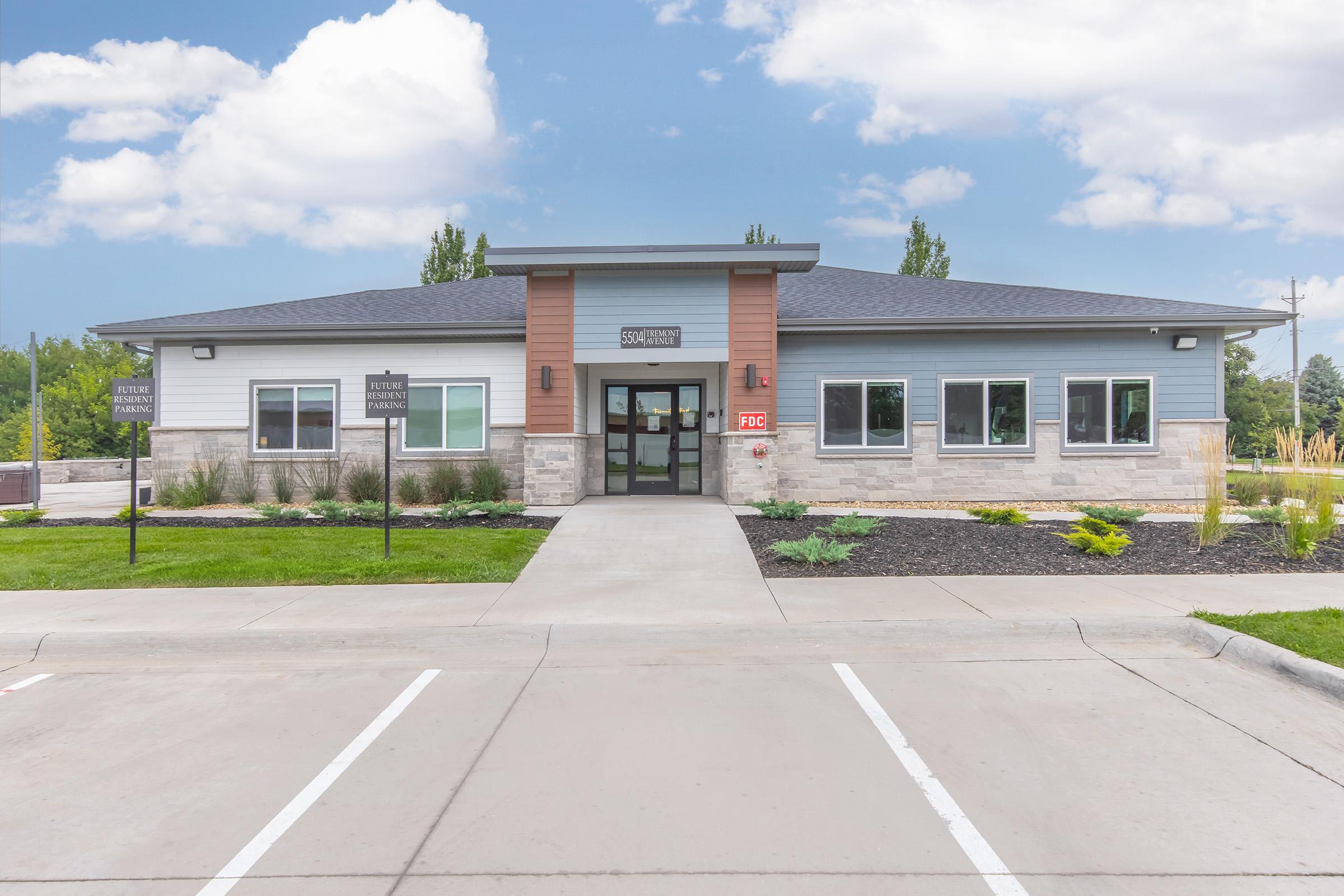 Modern single-story building with a combination of light gray and blue siding. Features large windows, a well-maintained front lawn, and a paved parking area. Signage indicates it is a business or community center, with a welcoming entrance and landscaped surroundings.