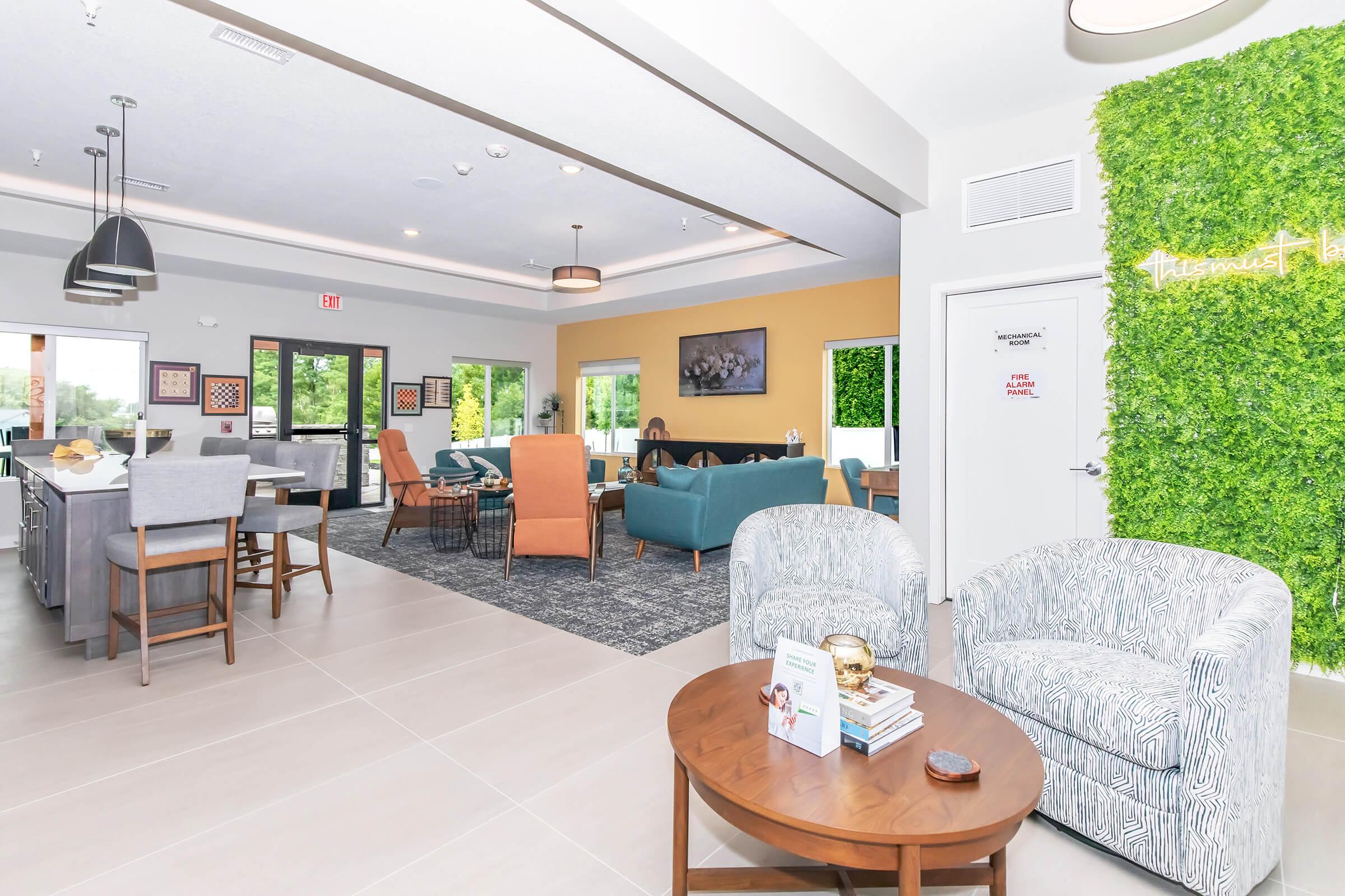 A modern interior space featuring a cozy lounge area with comfortable seating, including armchairs and a sofa. There are tables with books and decorative items, along with greenery on the wall. Large windows allow natural light to fill the room, creating a welcoming atmosphere.