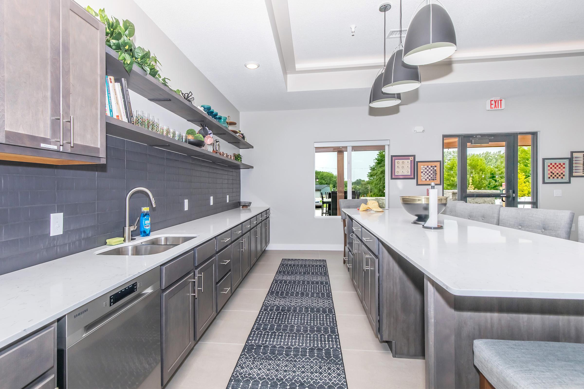 Modern kitchen featuring stainless steel appliances, dark cabinetry, and a long island with stools. The space includes a wall of shelves with decor, a sink, and a large window offering a view of greenery outside. The floor is tiled, and the overall design is sleek and contemporary.
