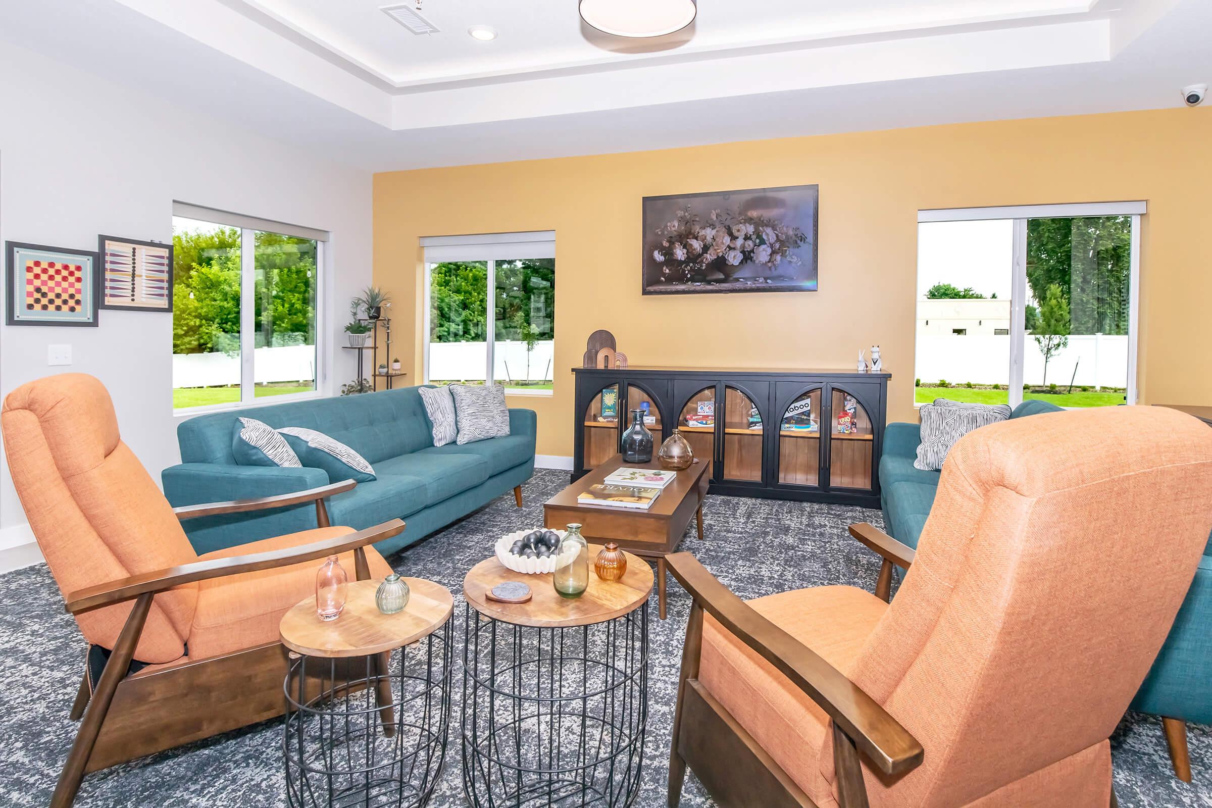 A cozy living room featuring two teal sofas and two orange armchairs arranged around a wooden coffee table. Large windows provide abundant natural light, showcasing a pleasant outdoor view. A flat-screen TV is mounted on the wall above a decorative cabinet, with artwork and plants adding warmth to the space.