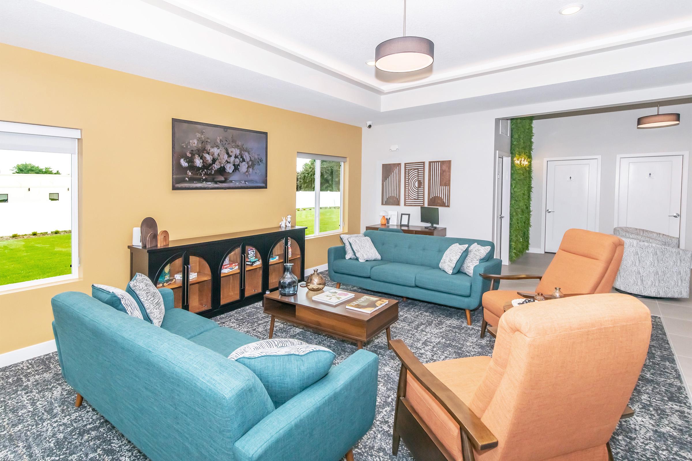 A modern living room featuring two cozy turquoise sofas and an orange armchair. There's a wooden coffee table in the center with decorative items, and a TV mounted on the wall. The room has large windows allowing natural light and a view of greenery outside, with a stylish rug and artwork adding to the decor.