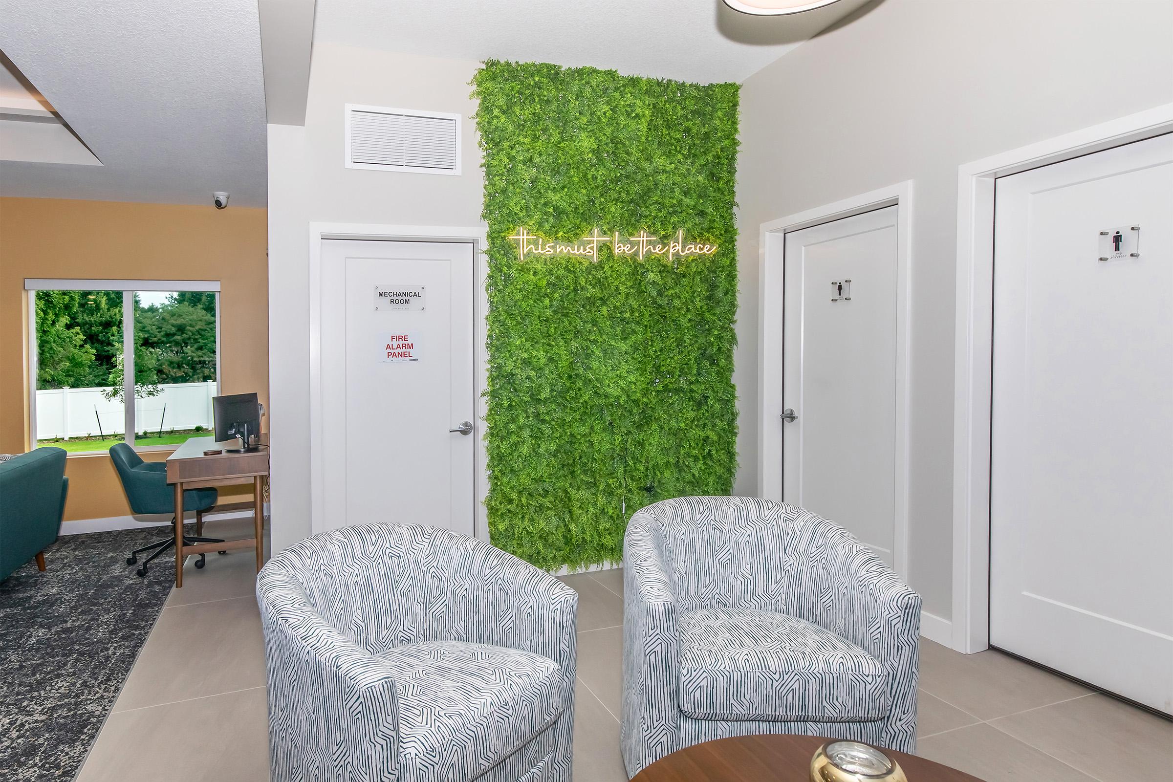 A modern lounge area featuring two patterned chairs facing a small round table. A wall covered in green foliage decor serves as a backdrop, with a light sign that reads "The Mints the Place." Doors labeled "Restroom" are visible nearby, along with a workspace area in the background.