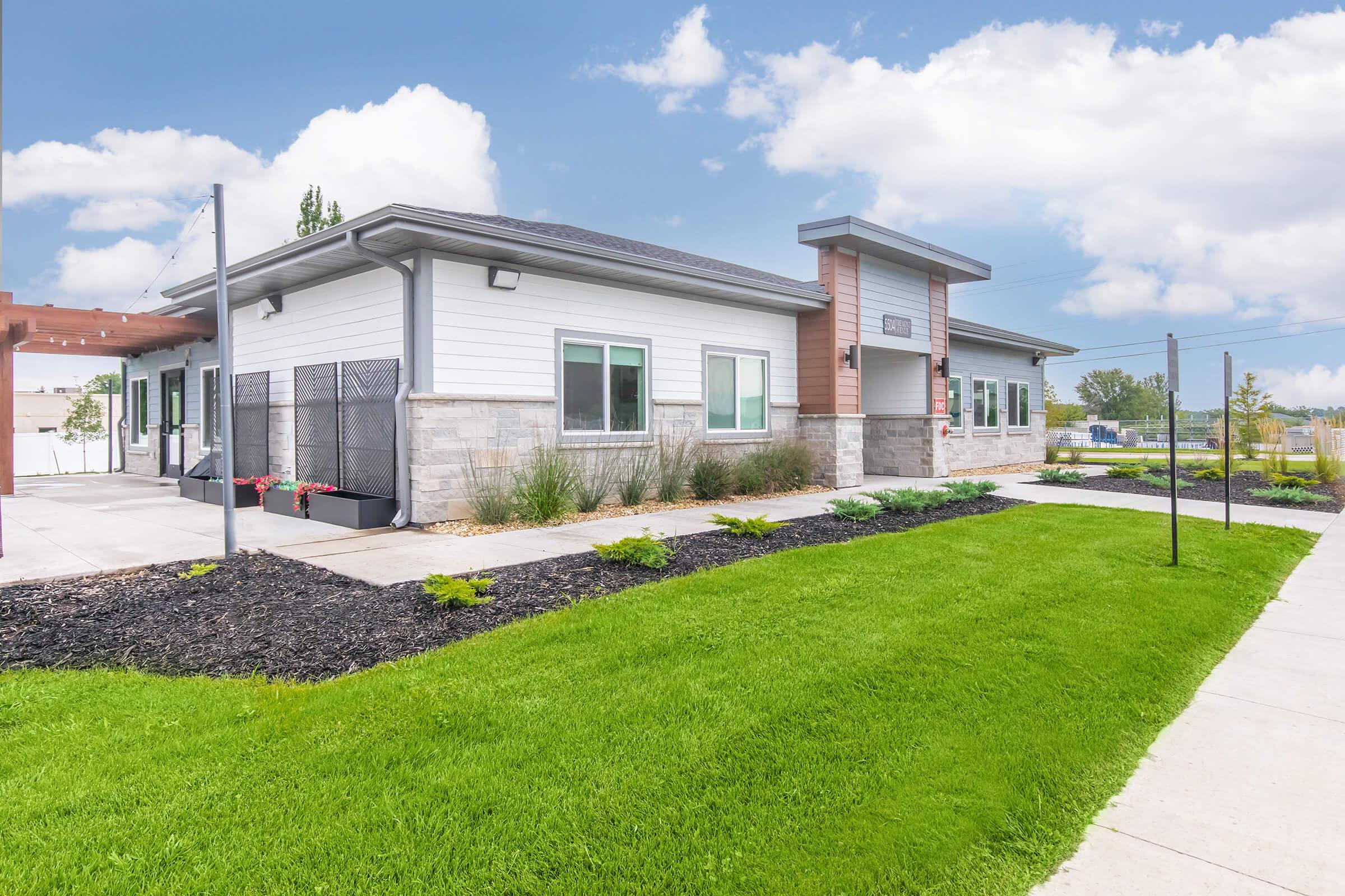 A modern single-story building with a sleek design, featuring a light-colored facade, large windows, and a welcoming entrance. The surrounding area includes manicured green lawns, neatly arranged flower beds, and a clear blue sky in the background, creating a bright and inviting atmosphere.
