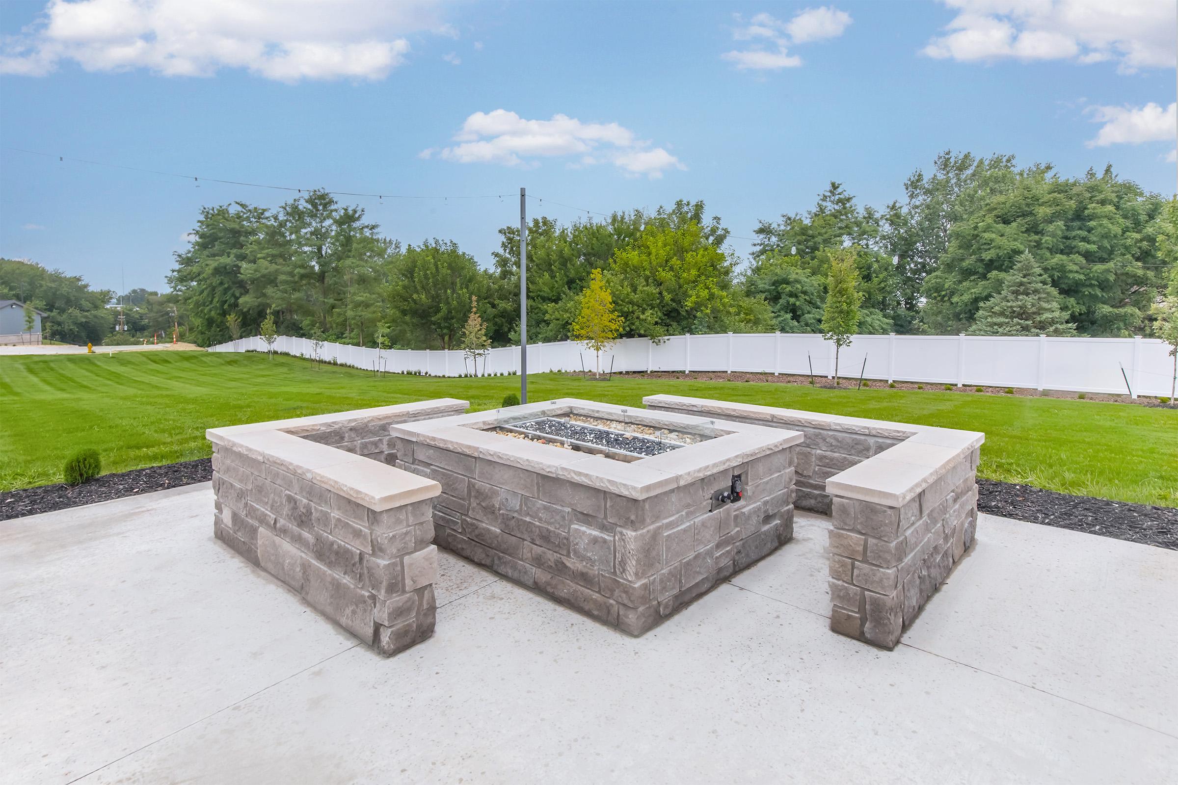 A landscaped outdoor area featuring a rectangular stone fire pit surrounded by low stone walls. Lush green grass and trees line the background, with a white fence visible. The sky is bright with a few clouds, indicating a clear day.