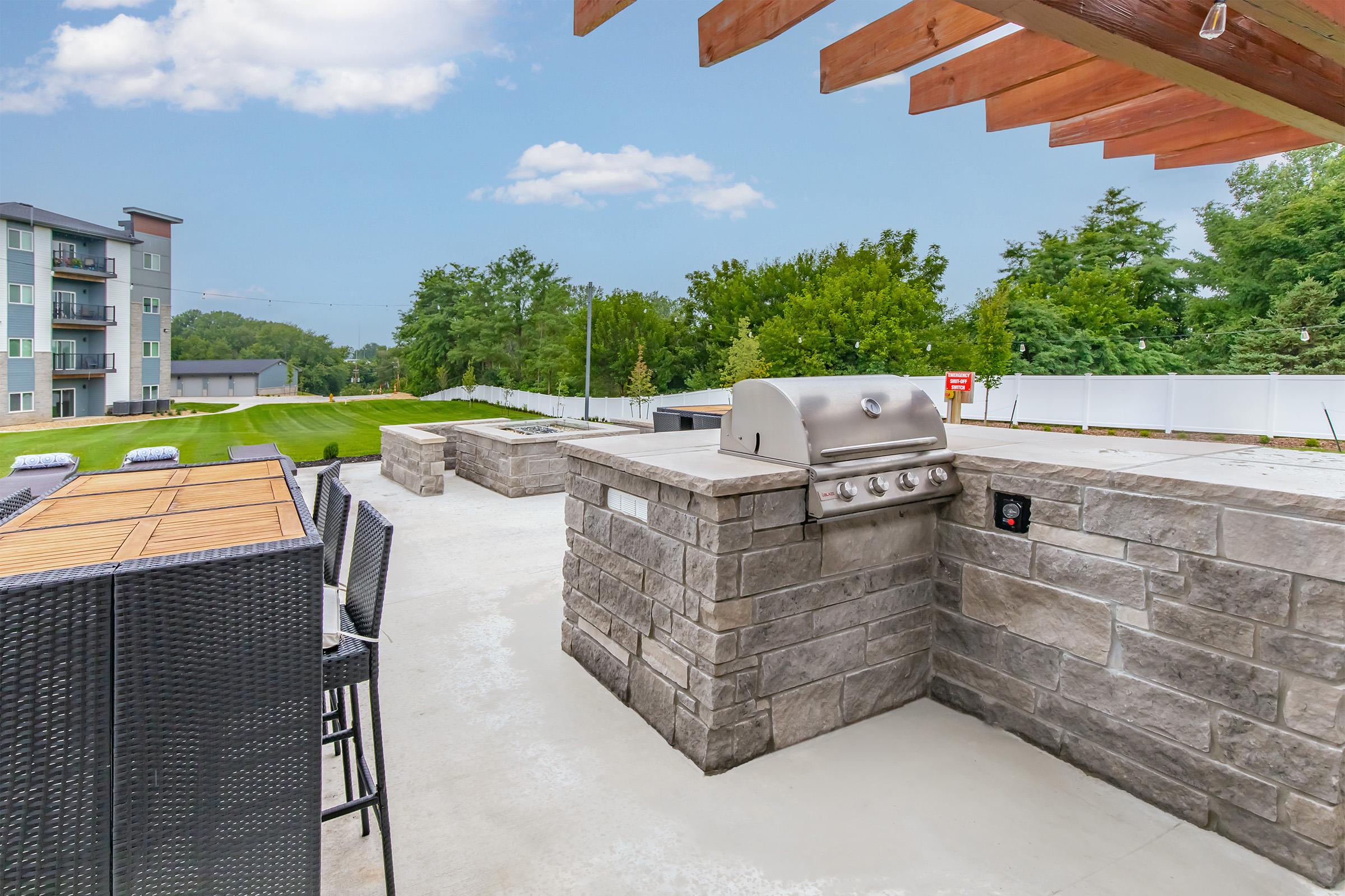 Outdoor grill area with a built-in stone barbecue, bar-style seating nearby, and a view of a green lawn and trees. The setting is well-lit and features a clear blue sky with some clouds, creating a welcoming atmosphere for gatherings or cooking outdoors.
