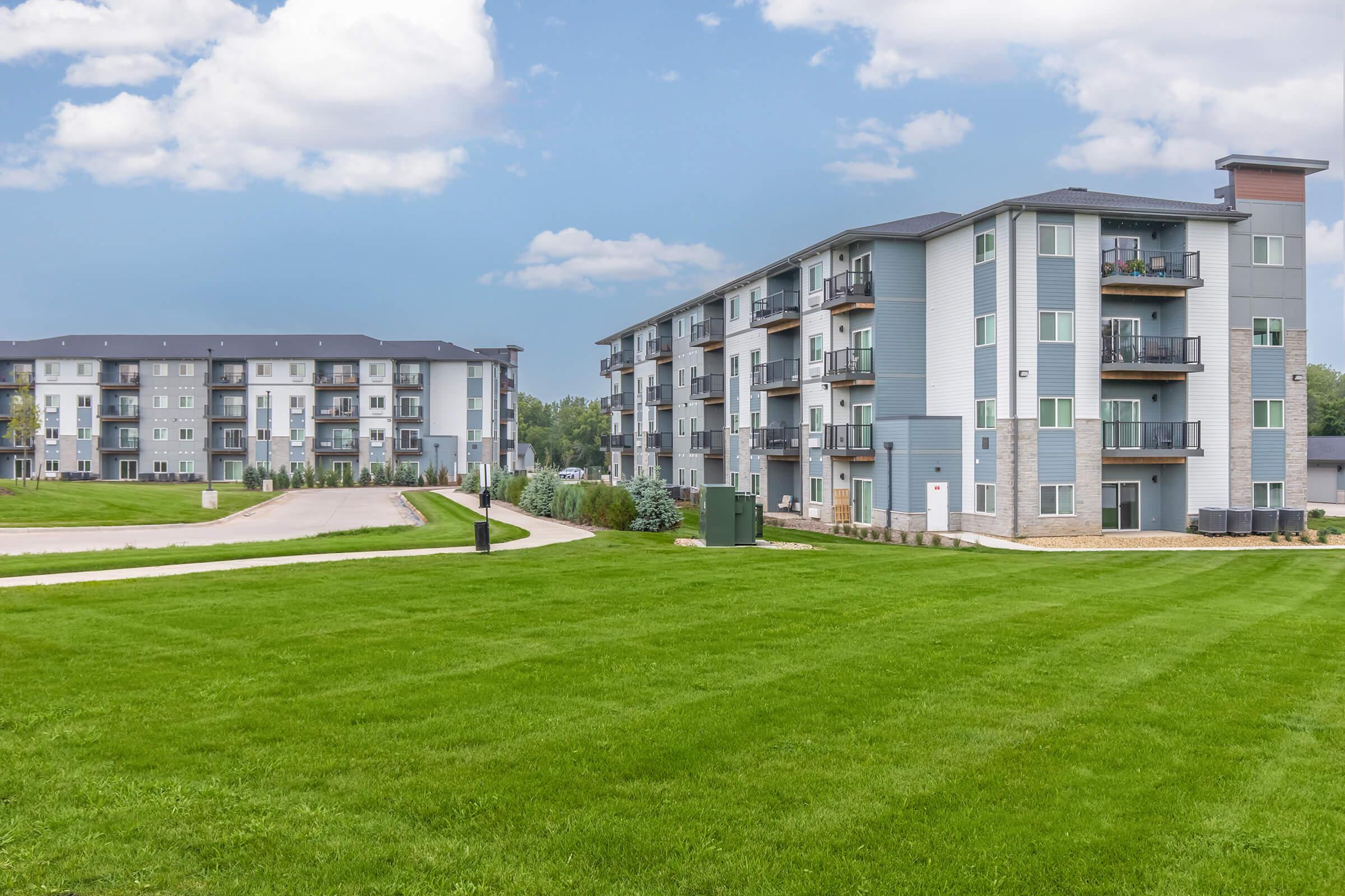 A wide view of a modern residential complex featuring two three-story apartment buildings. The buildings are surrounded by well-maintained green lawns and a pathway. The sky is partly cloudy, creating a bright and inviting atmosphere. Ideal for showcasing contemporary living spaces.