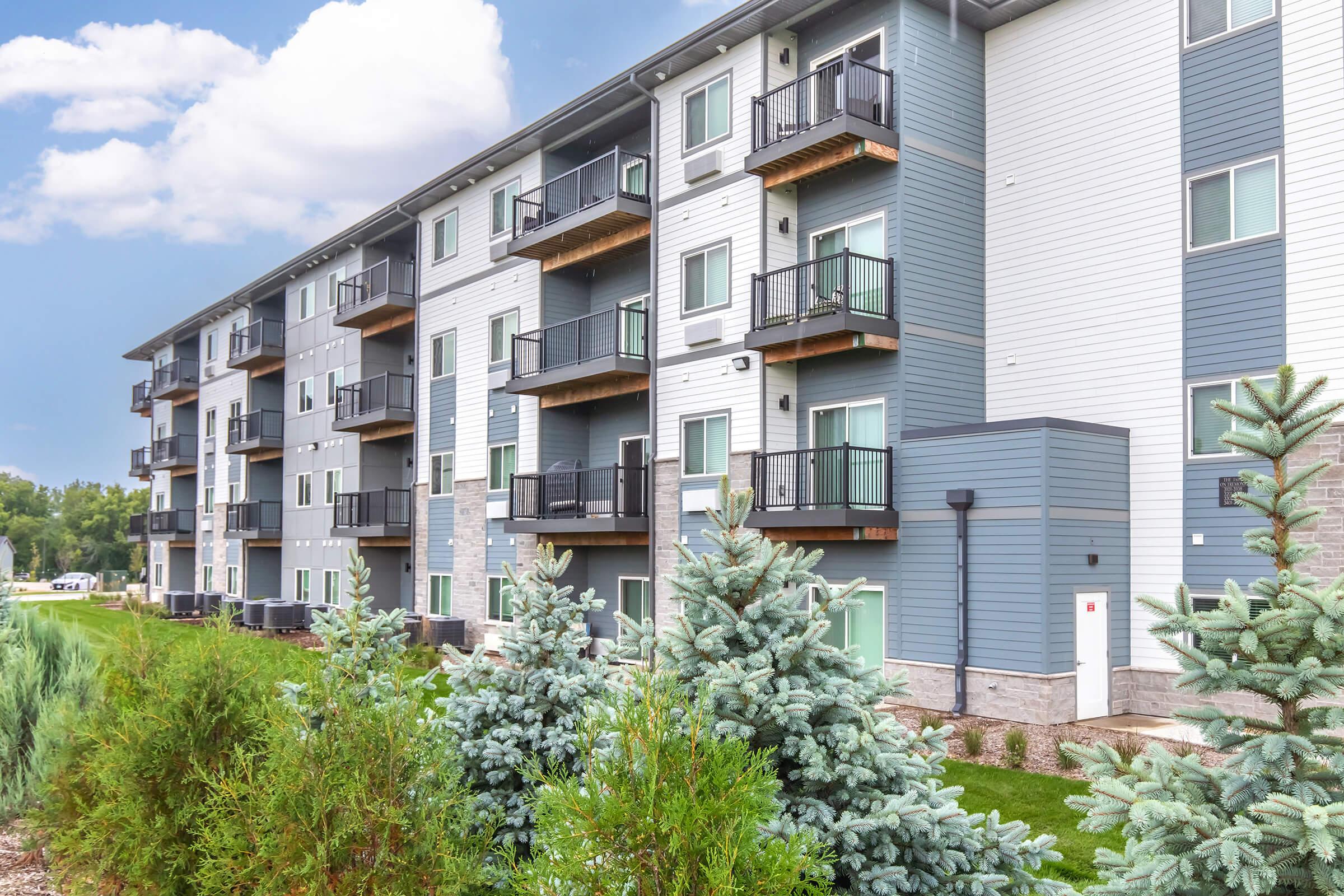 Modern multi-story residential building with balconies, featuring a mix of light and dark siding. In the foreground, there are evergreen shrubs and trees, while the background includes a clear blue sky with a few clouds. The setting appears landscaped and well-maintained.