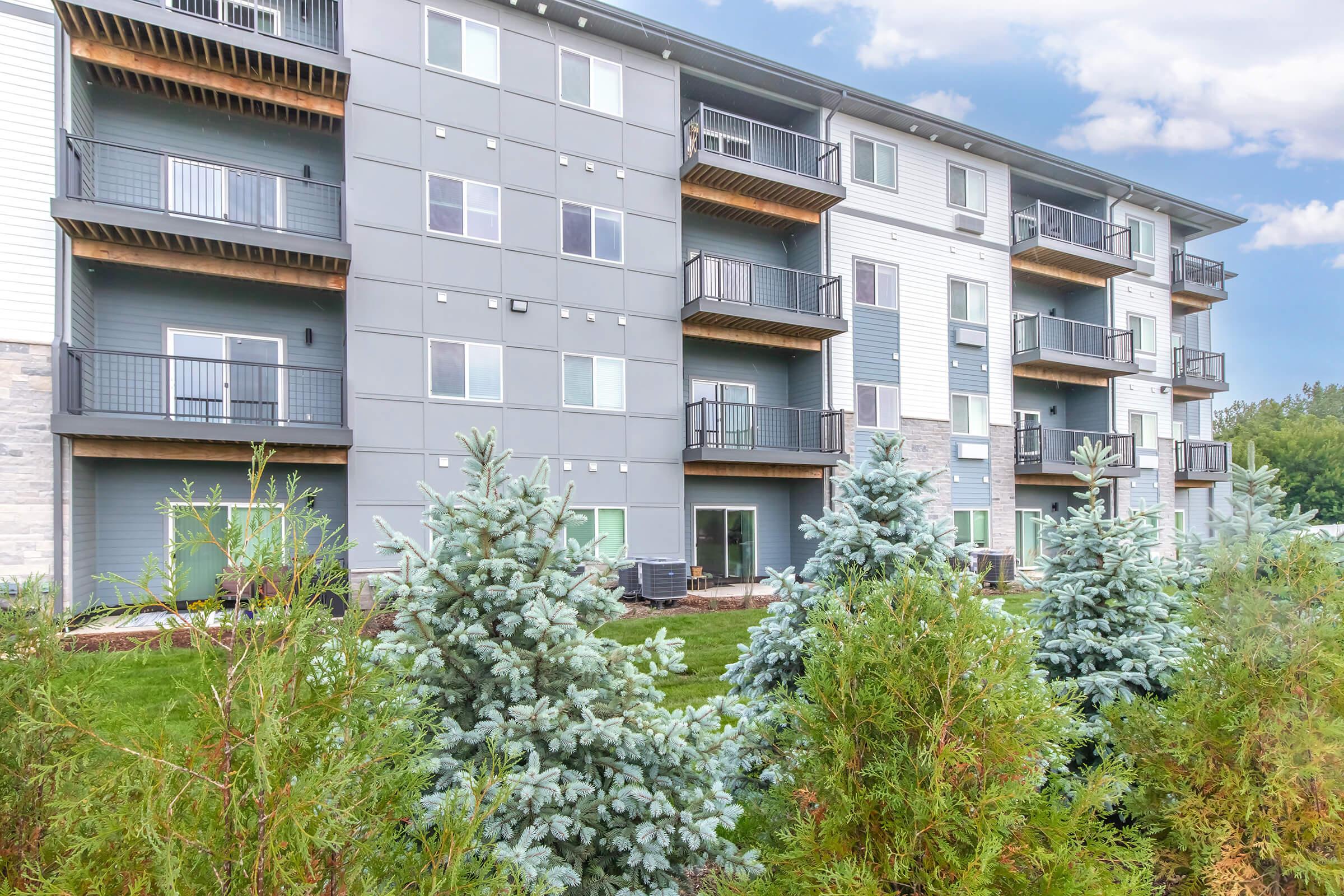 A modern apartment building with multiple floors and balconies. The exterior features a combination of gray and blue siding. In the foreground, there are neatly trimmed evergreen shrubs, and the setting includes a well-maintained grassy area under a partly cloudy sky.