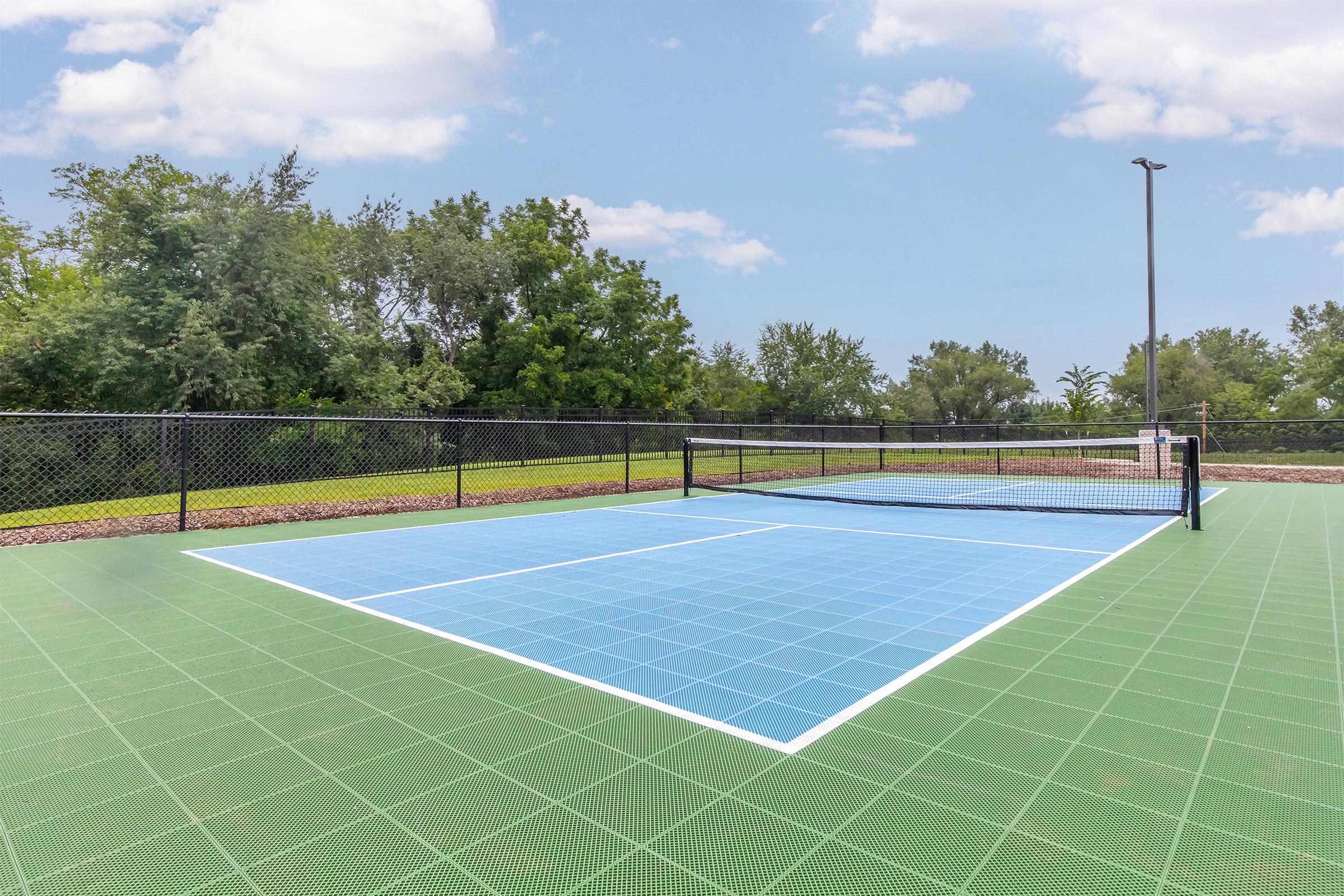 A well-maintained outdoor tennis court featuring a light blue playing surface, surrounded by a black fence and lush greenery in the background. The sky is partly cloudy, providing a bright atmosphere for playing tennis.