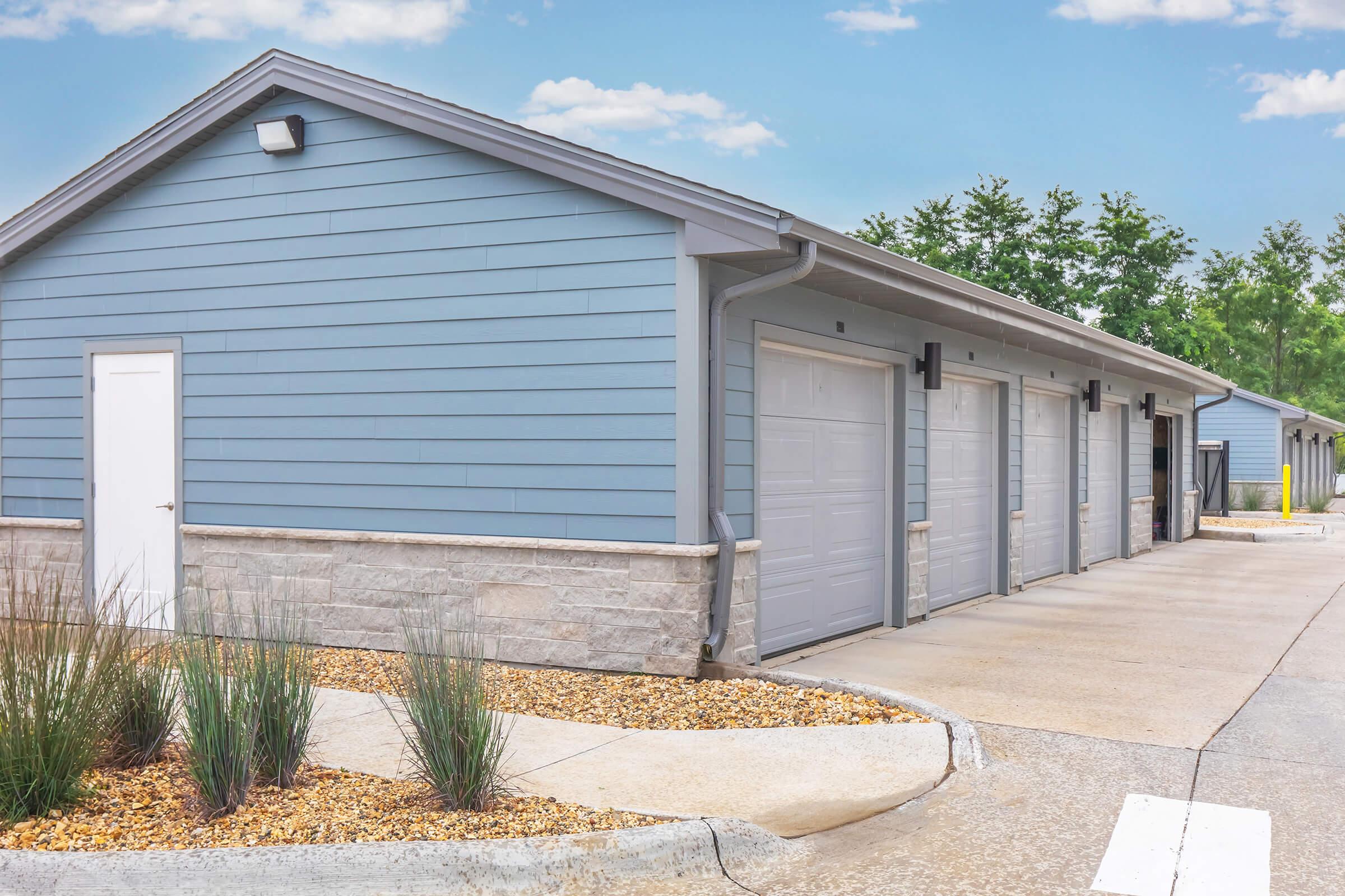 A modern, single-story building with light blue siding and stone accents, featuring multiple garage doors. The pathway in front is paved and bordered by gravel and small plants. In the background, greenery and a blue sky are visible, creating a serene atmosphere.