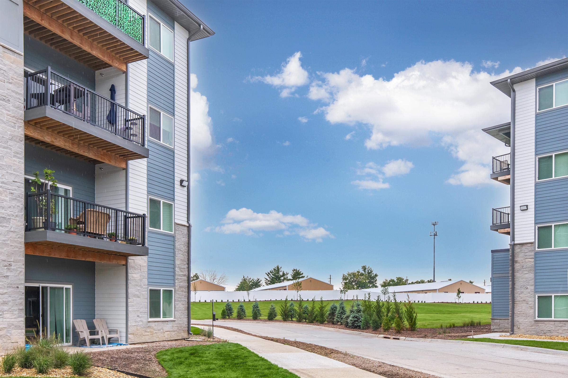 A residential area featuring modern apartment buildings with balconies, situated alongside a paved walkway. The sky is clear with a few clouds, and there are manicured lawns and trees in the background, suggesting a pleasant outdoor environment.