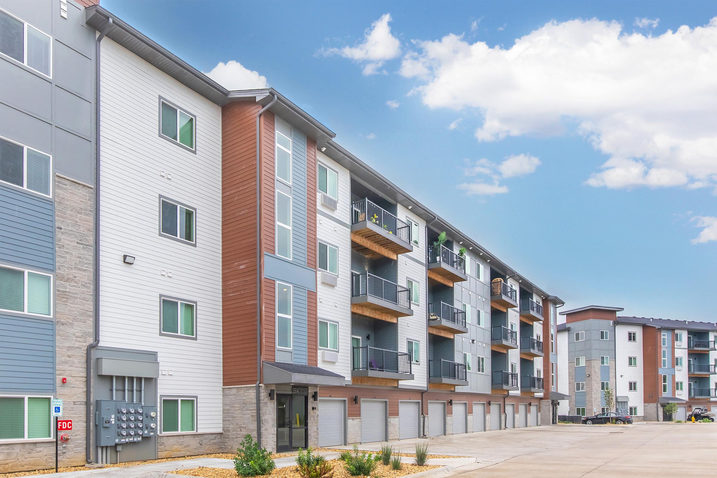 Modern apartment building with a mix of light gray and brown siding. The three-story structure features balconies on the upper floors, large windows, and a row of mailboxes at the entrance. The scene includes a clear blue sky with a few clouds, and a paved area in front with some landscaping.