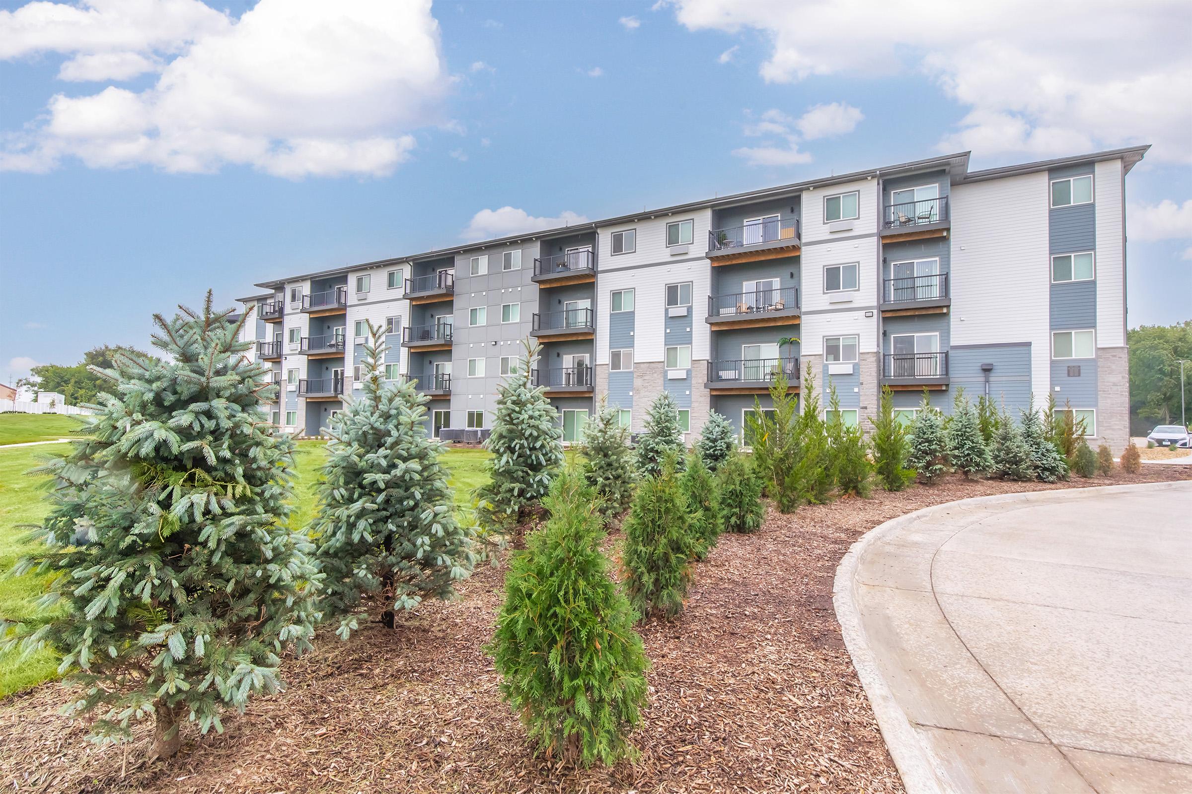A modern, multi-story apartment building surrounded by well-maintained landscaping. In the foreground, there are neatly arranged coniferous trees and shrubs, while the building features balconies and large windows. The sky is partly cloudy, adding a pleasant backdrop to the scene.
