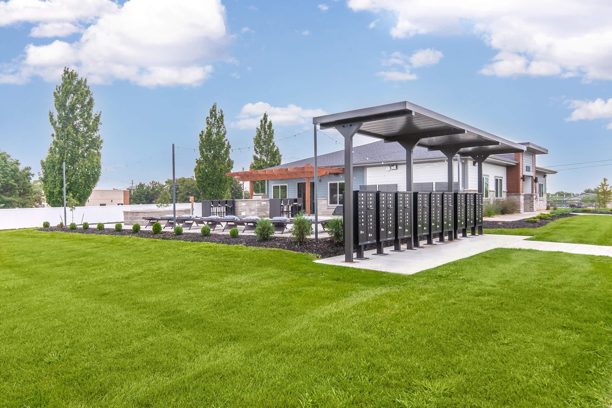 A well-maintained outdoor area featuring a green lawn, modern mailboxes on the left, and a shaded seating area with lounge chairs. In the background, there are buildings with a mix of architectural styles under a clear blue sky, creating a pleasant community atmosphere.