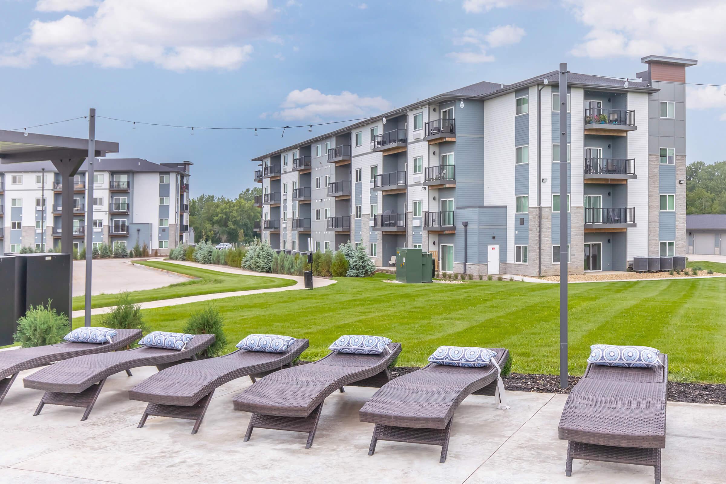 A view of a modern apartment complex featuring multiple buildings in the background, with well-maintained grassy areas. In the foreground, there are several stylish lounge chairs arranged neatly on a patio, creating a relaxing outdoor space. The sky is partly cloudy.