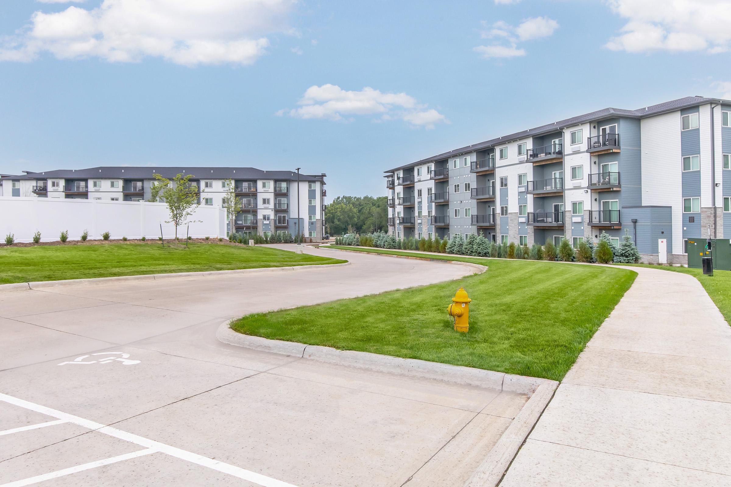 A view of a modern apartment complex with multiple buildings, surrounded by well-maintained green lawns. A curved driveway leads through the area, with a yellow fire hydrant visible on the grass. The sky is clear and blue, adding to the fresh and inviting atmosphere of the residential community.
