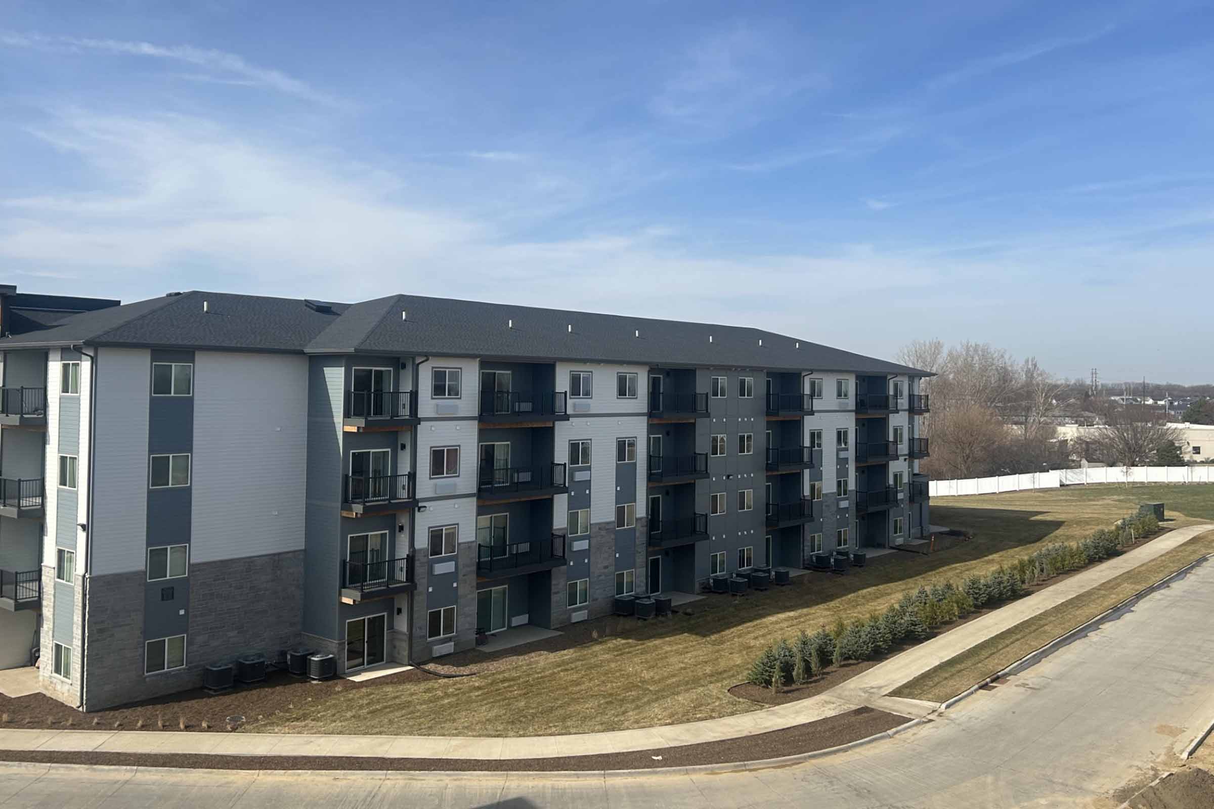 A modern multi-story apartment building with a mix of gray siding and stone accents, featuring balconies on the upper floors. In the foreground, there's a well-maintained lawn with shrubs and a curved pathway leading to the street. The sky is blue with a few clouds visible.