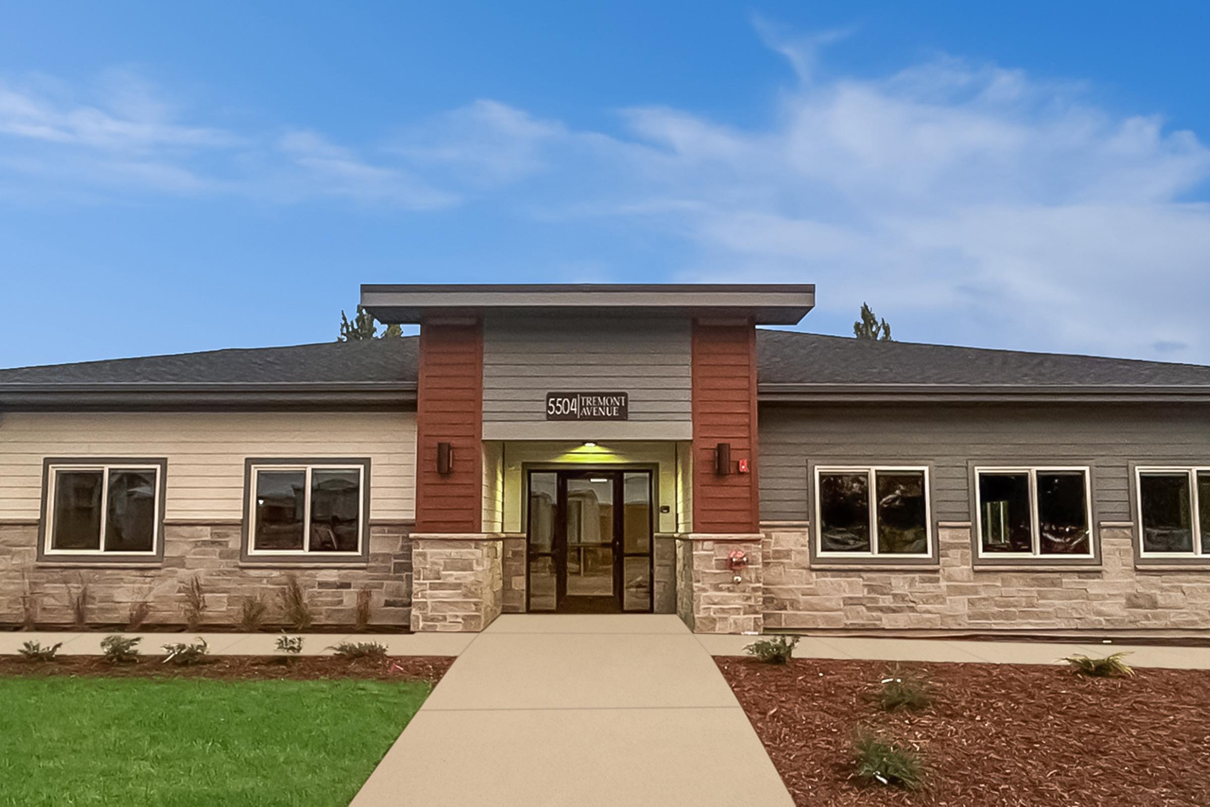 A modern building with a combination of light-colored siding and stone accents. The entrance features a covered porch, large windows, and a sign displaying the address "5504 Hermans Avenue." The surrounding area has landscaped mulch and shrubs under a clear blue sky.