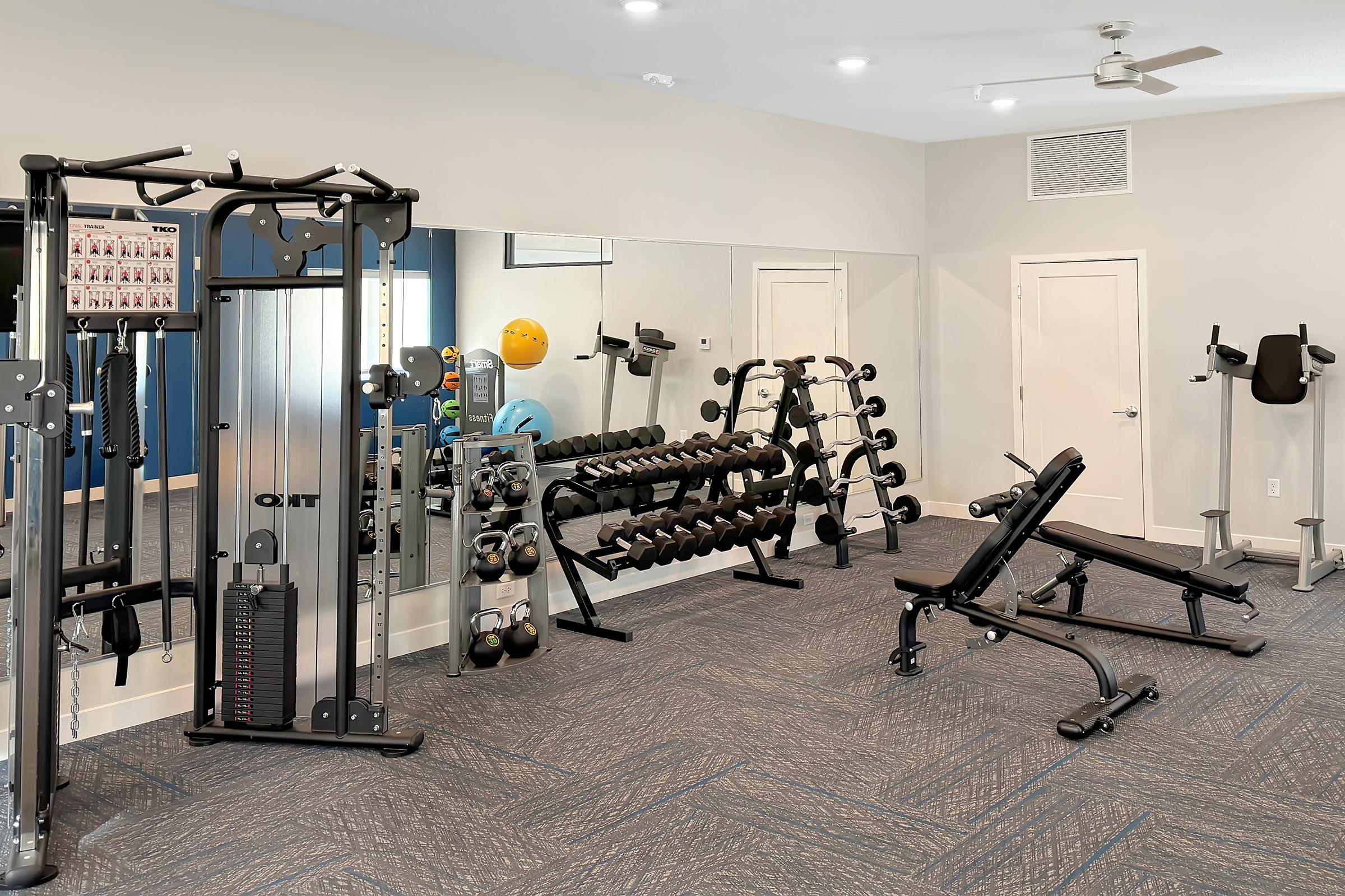 A modern gym interior featuring a mirror, various weightlifting equipment, including dumbbells on racks and a multi-station machine. There is also a bench for workouts and fitness balls nearby. The space is well-lit with a clean, organized layout.
