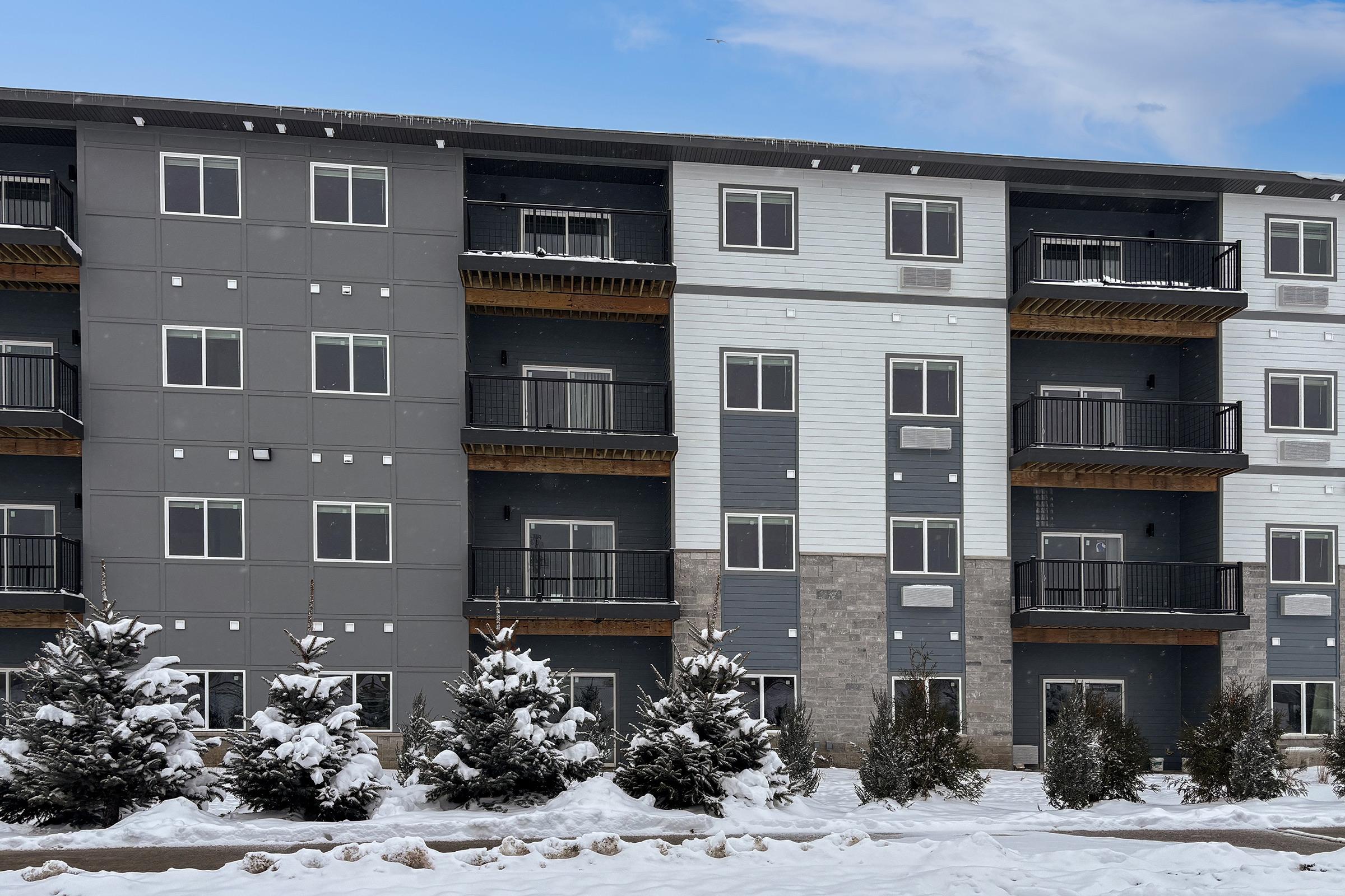 A modern multi-story apartment building with a mix of gray and white siding. The building features multiple balconies and is surrounded by snow-covered ground and small evergreen trees, indicating a winter setting. A clear blue sky is visible in the background.